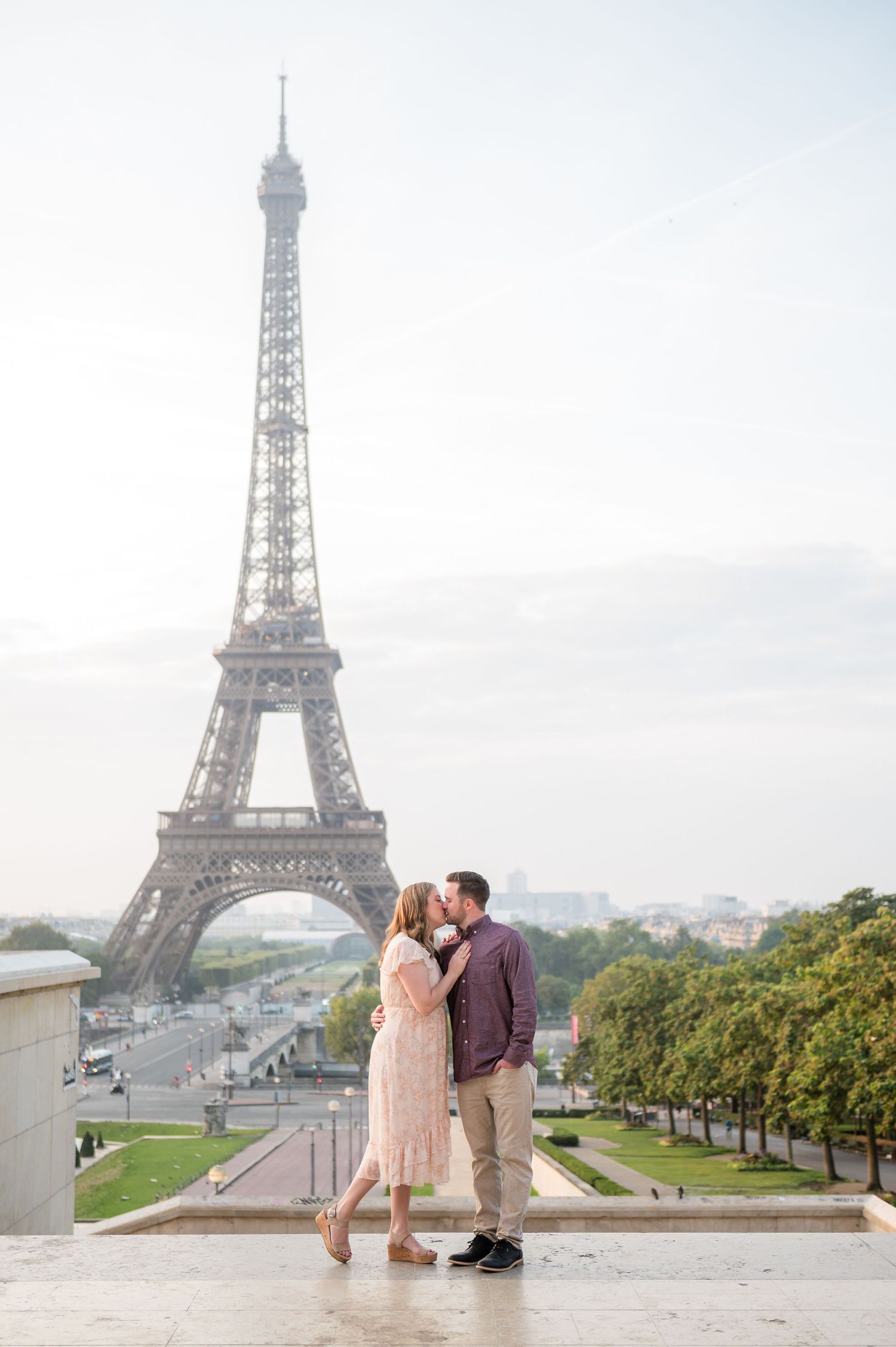 Couple in elegant attire embrace in front of Eiffel Tower on sunny Paris morning.