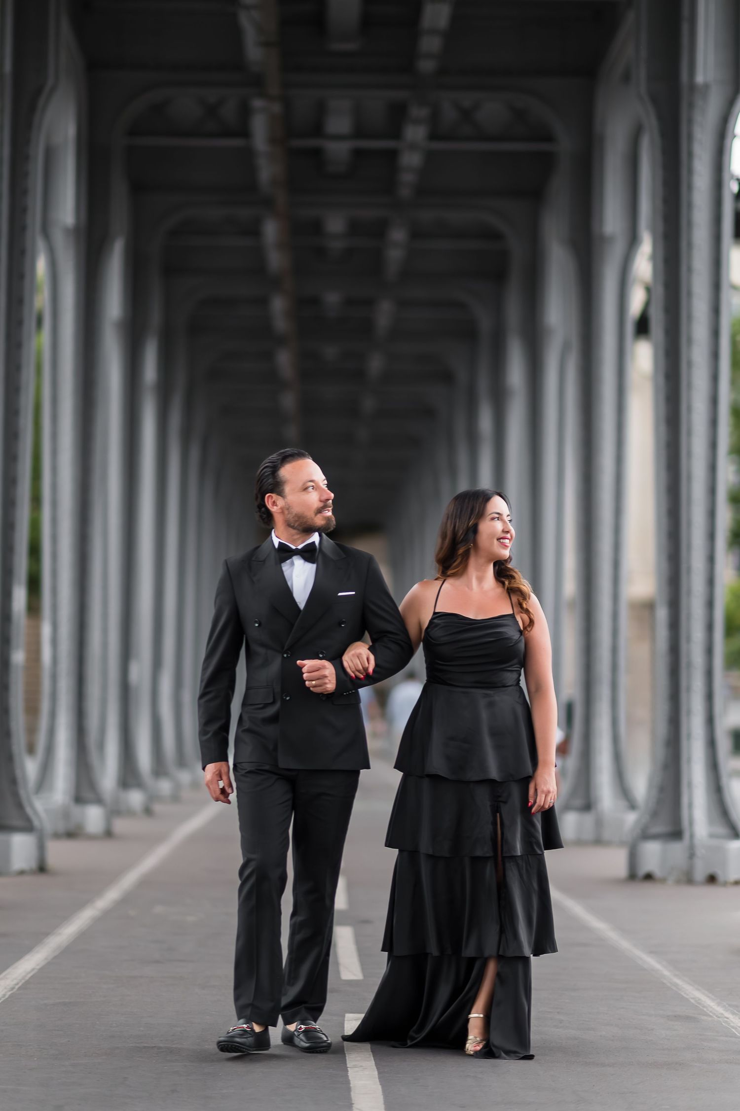 Elegant couple in matching black formal attire walking hand in hand under a grand bridge colonnade.
