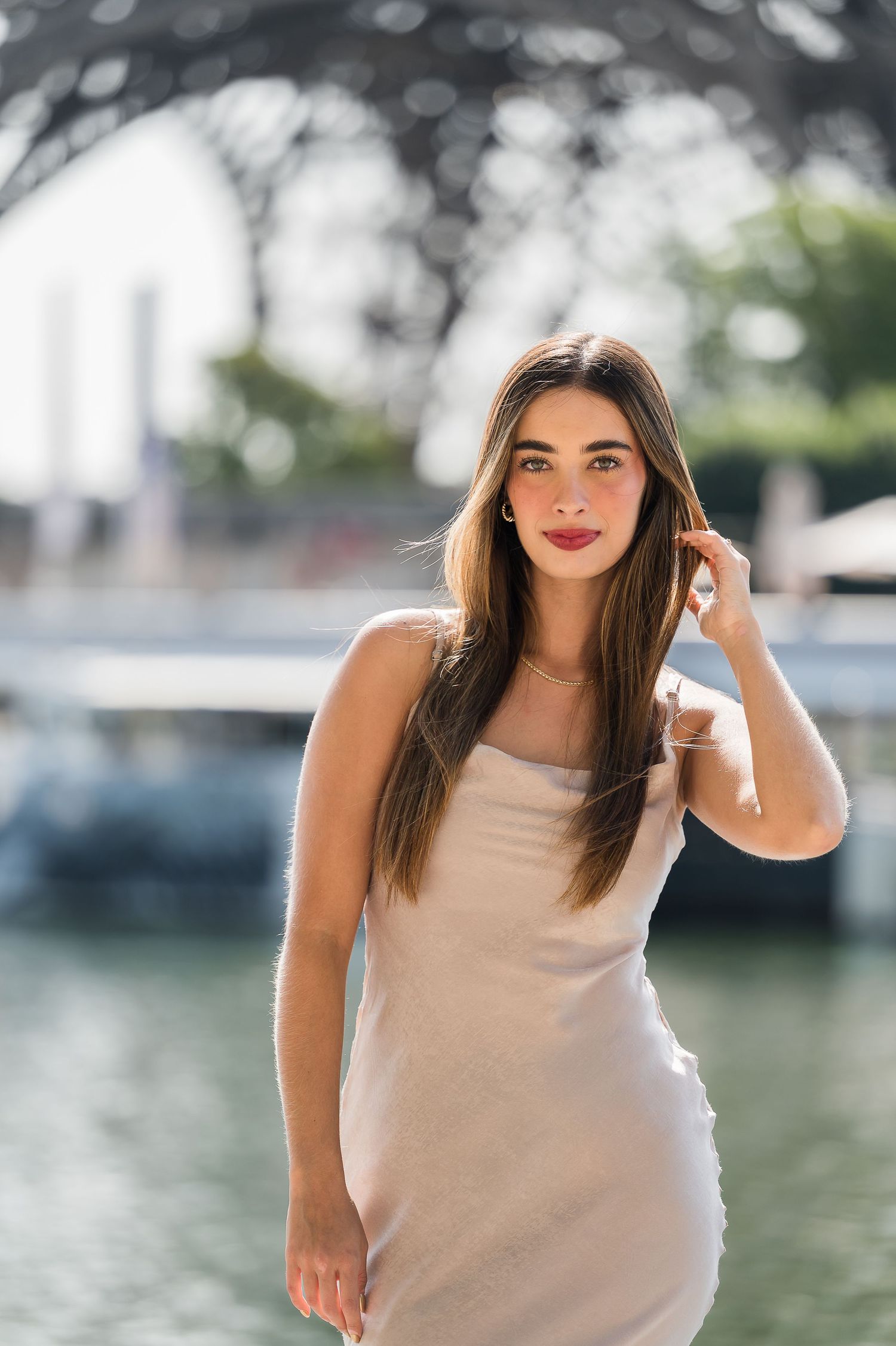 Young woman in white slip dress with long brown hair poses near a waterfront, possibly near the Eiffel Tower.