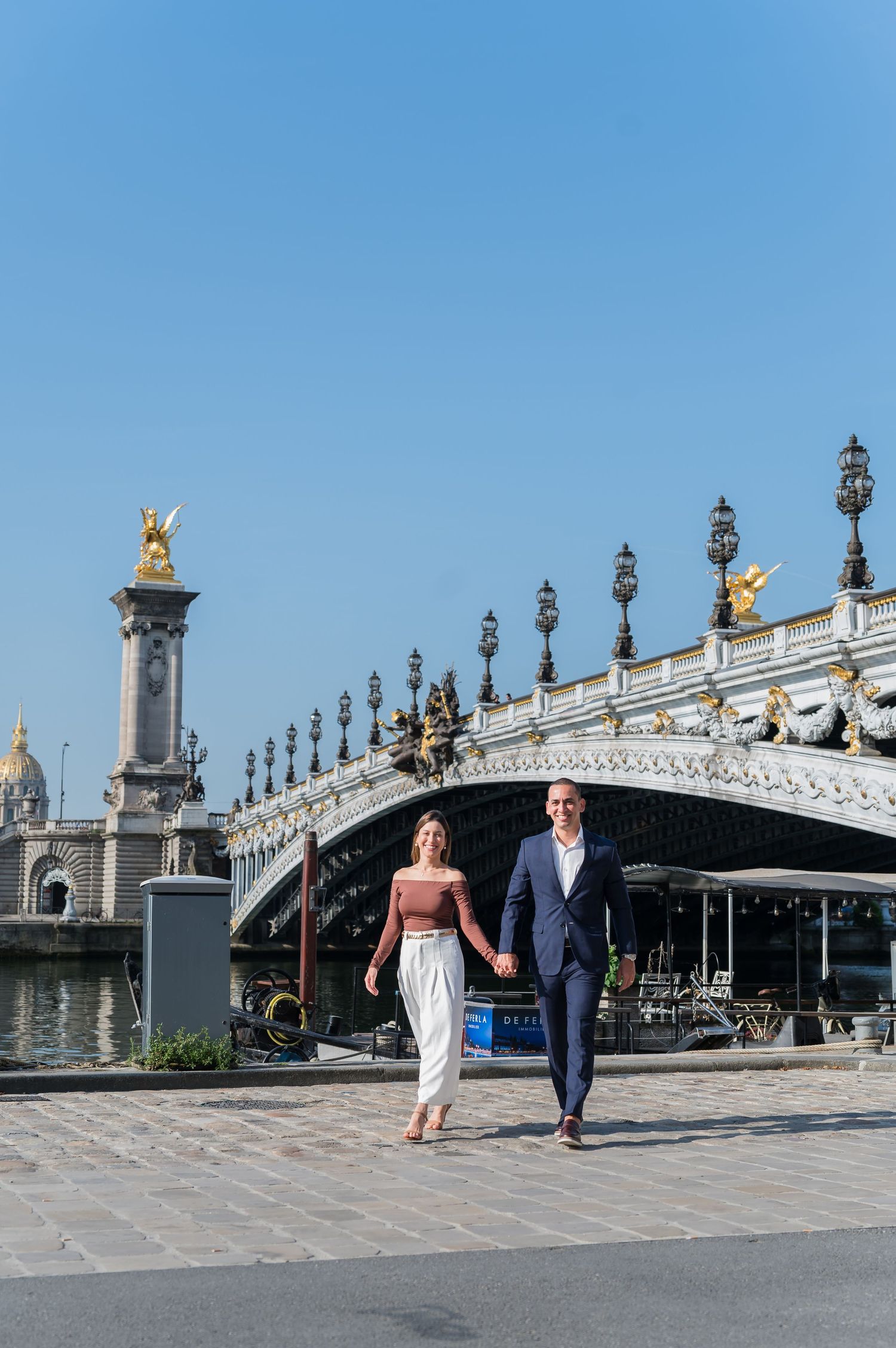 A romantic couple strolls along the iconic Pont Alexandre III bridge in Paris, known for its ornate golden statues and lamps.
