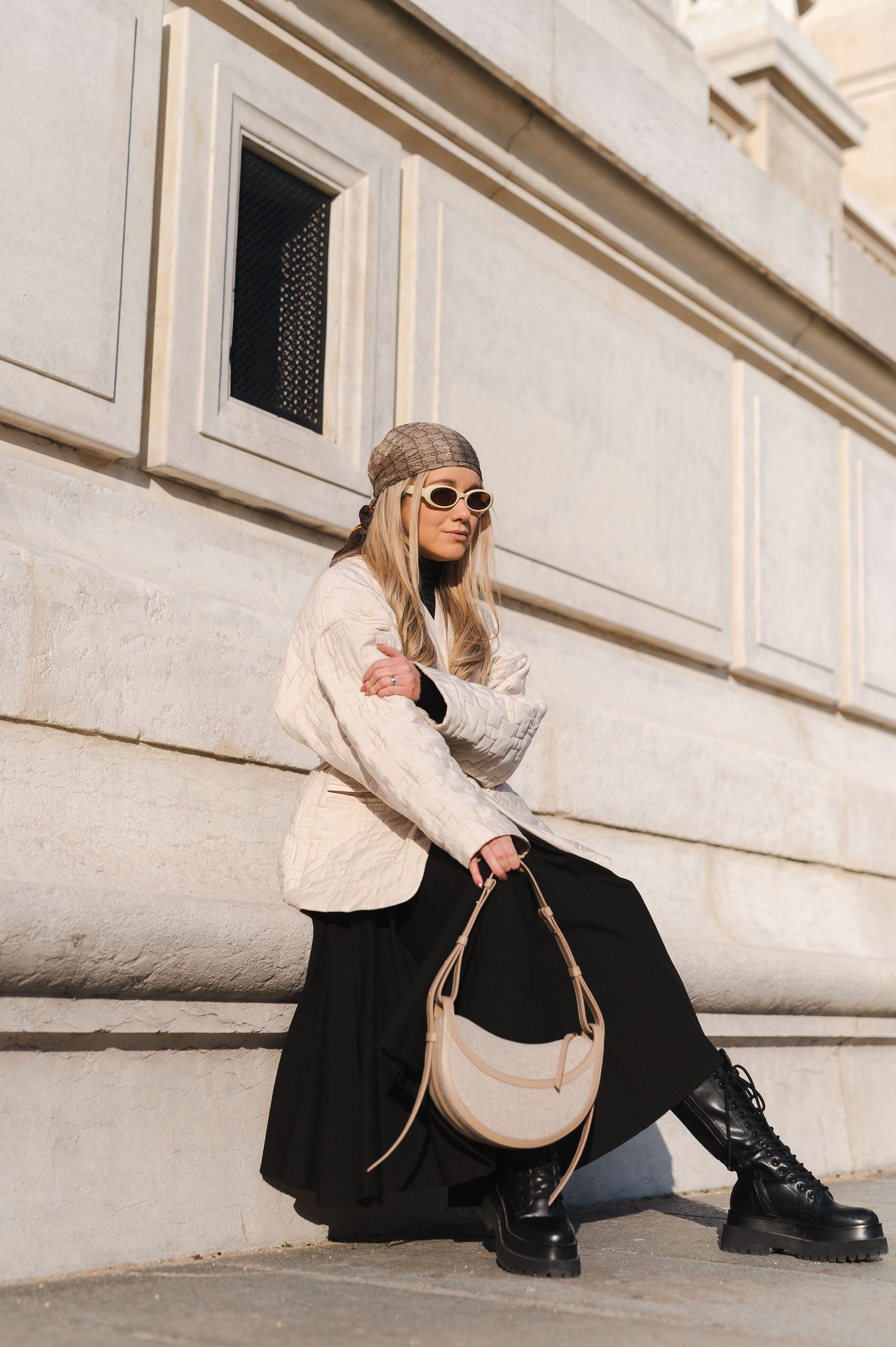 Fashion blogger in cream jacket and black skirt poses with handbag against stone building wall in sunlight.