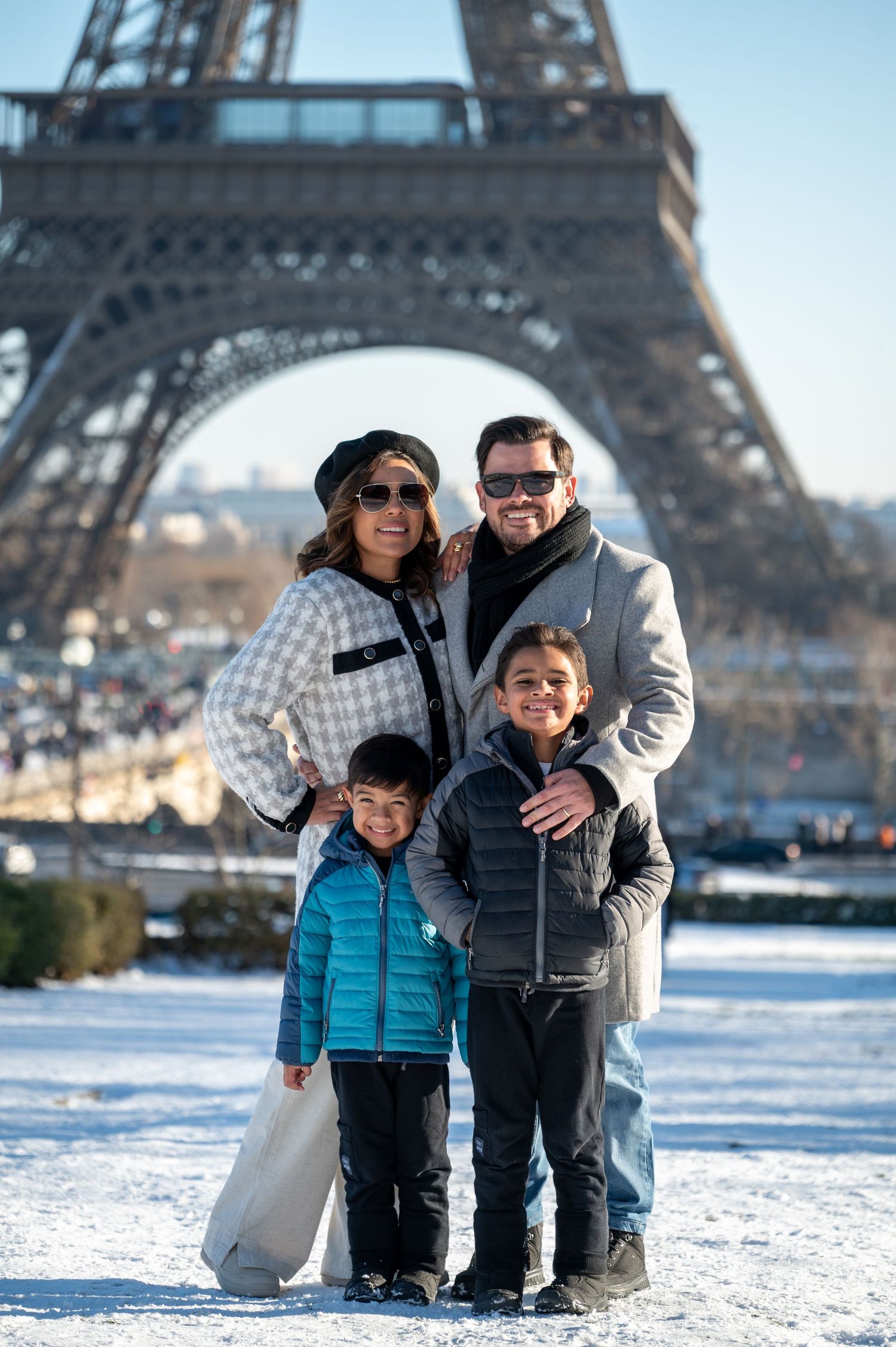 A family poses together in the snow with the iconic Eiffel Tower visible in the background on a winter day in Paris.
