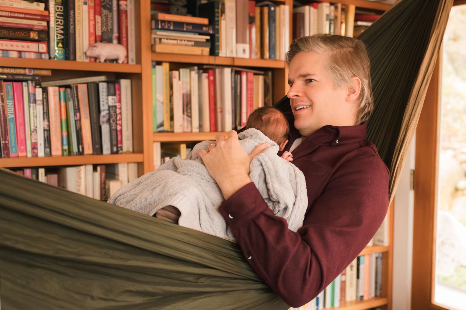 Person relaxing in a hammock holding a newborn baby in front of a large wooden bookshelf filled with books.