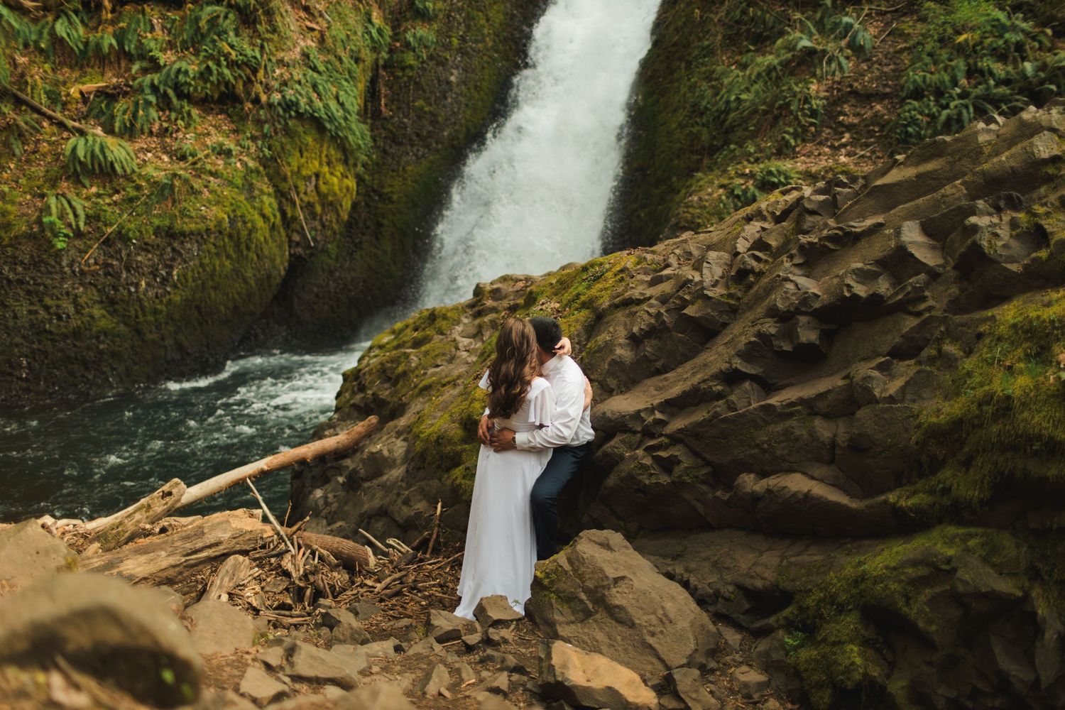 Couple embracing romantically on mossy rocks in front of a stunning waterfall in a lush green forest.