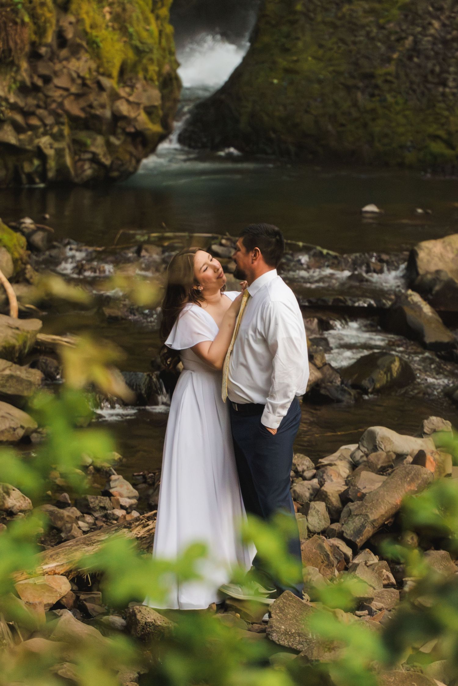 Couple in wedding attire embracing on rocky riverbank with waterfall and mossy cliffs in lush forest background.