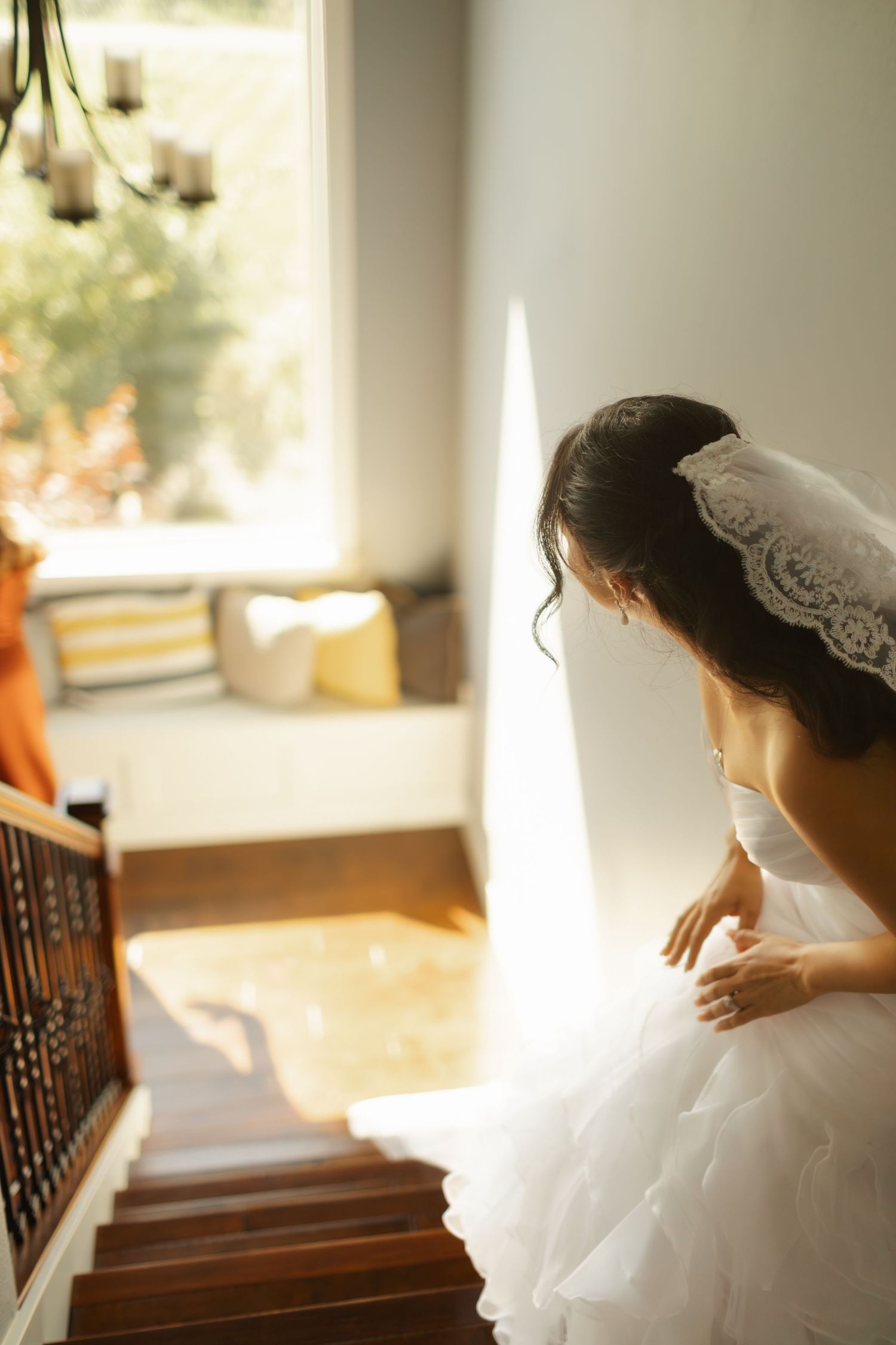 A bride in a white lace dress and veil gazes out of a sunlit window in a contemplative moment.