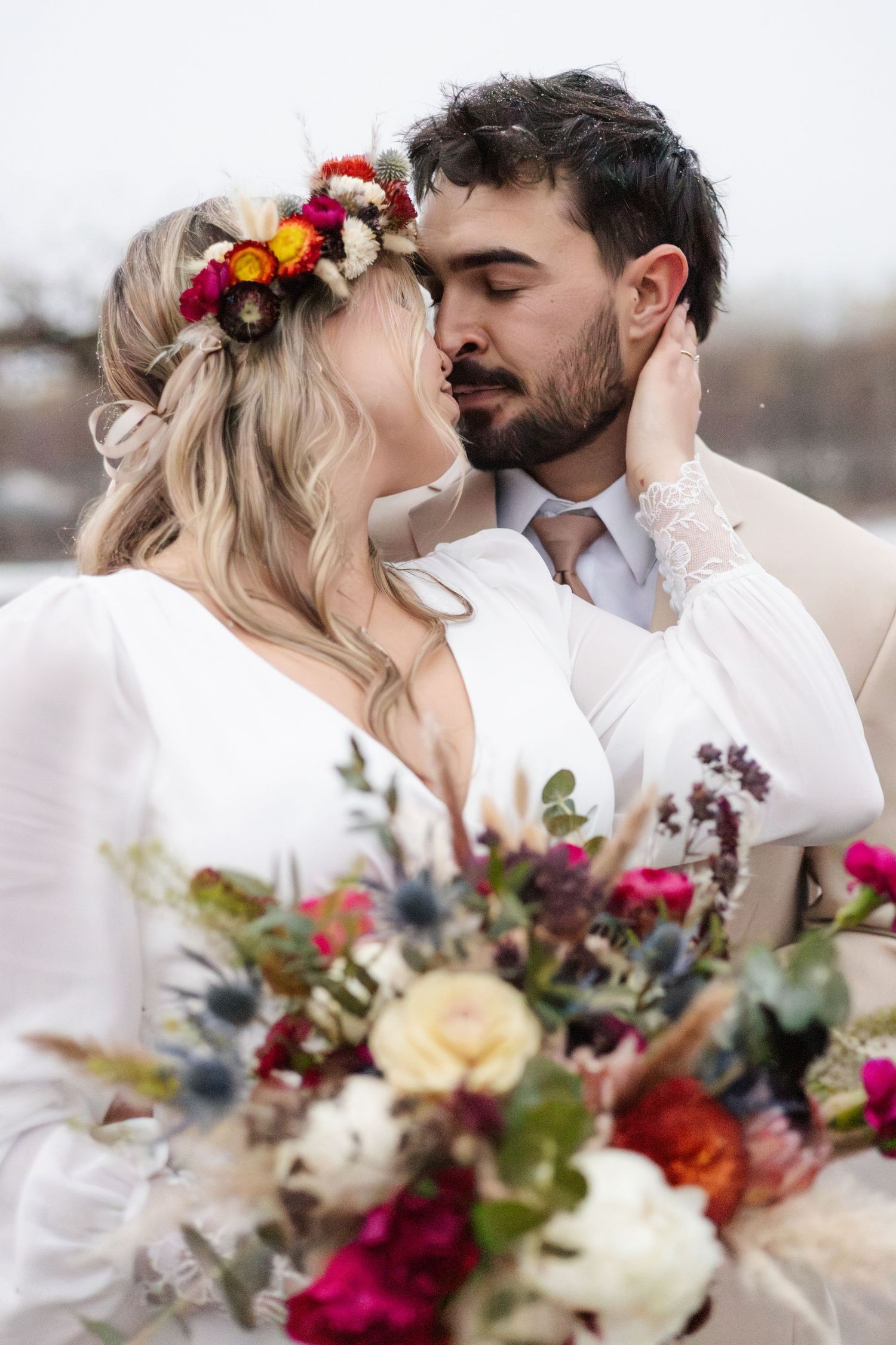 Bride and groom embrace in snowy setting, she in white dress with floral crown, he in tan suit.