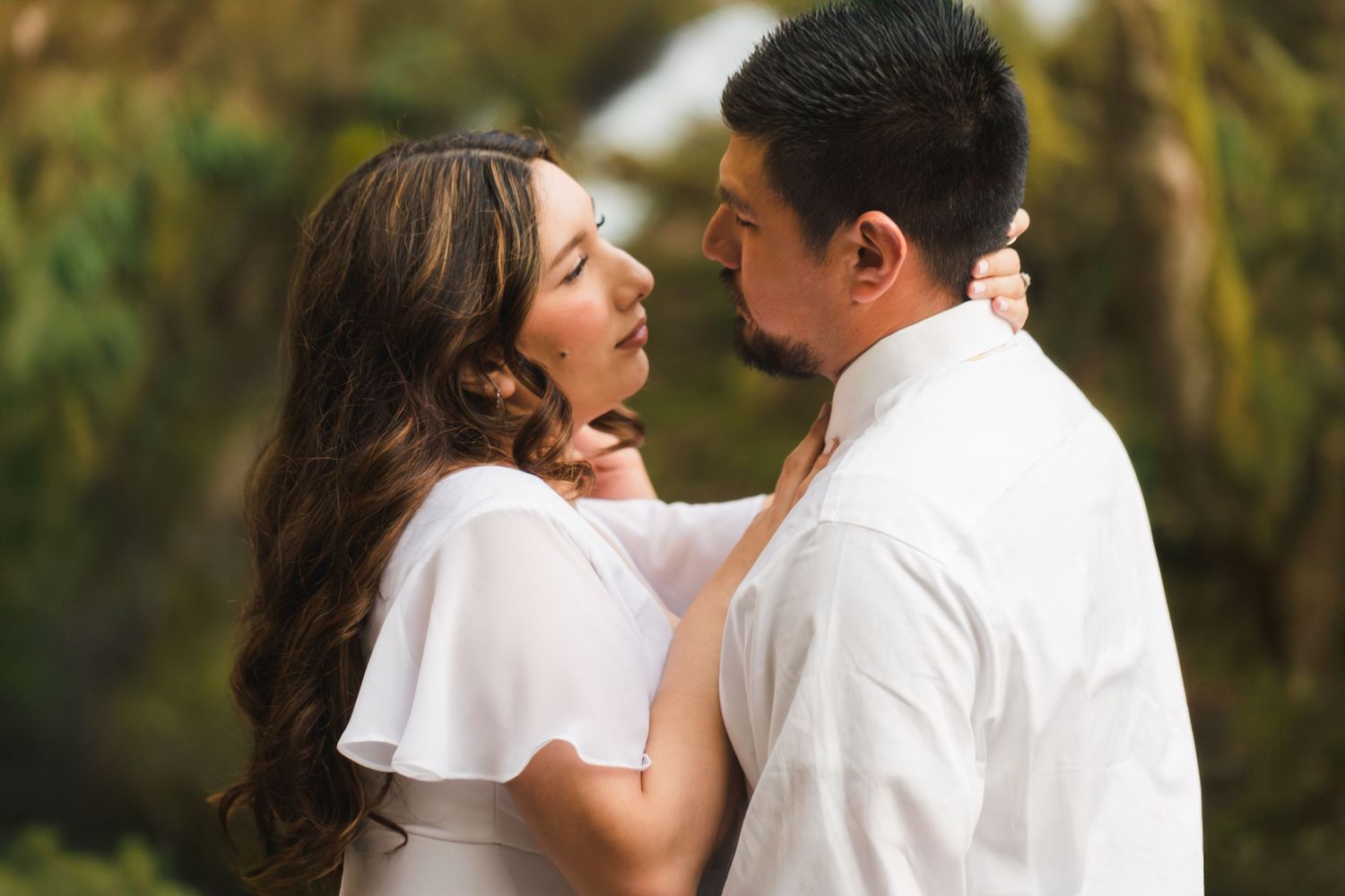 Romantic couple in white outfits sharing an intimate moment outdoors with lush greenery in the background.