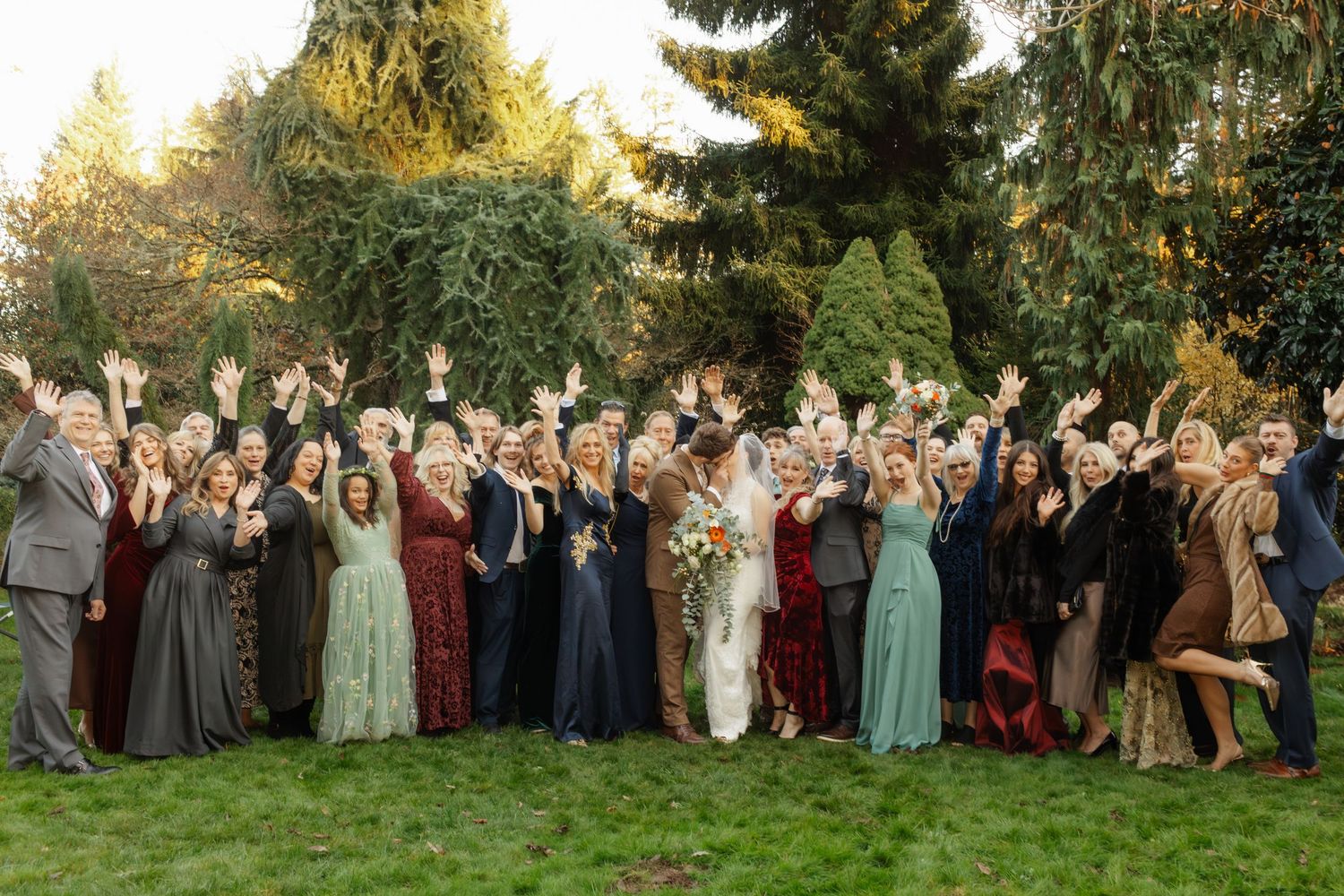Large wedding group photo with guests in mix of formal attire gathered outdoors at sunset.