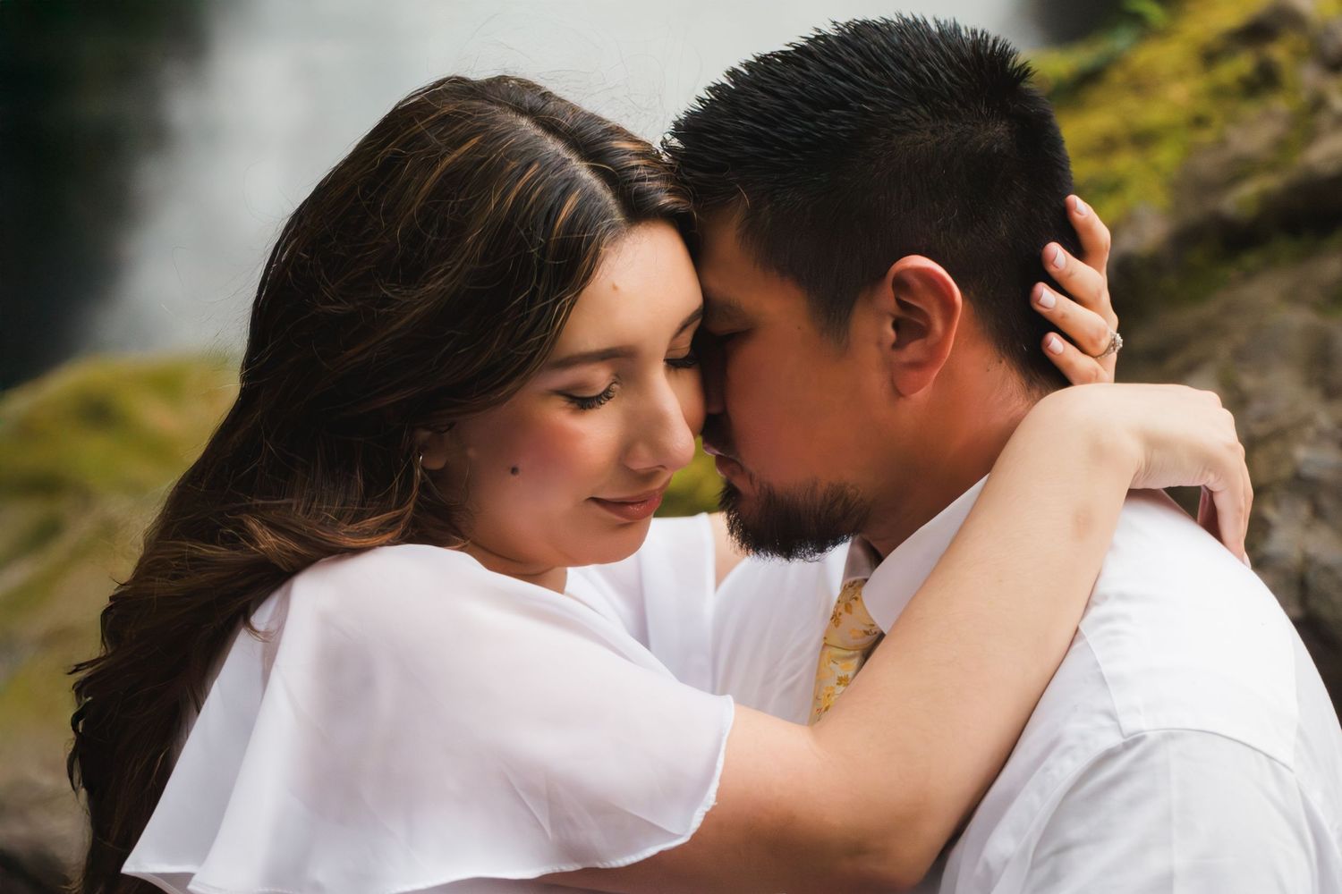 A couple in white attire share an intimate forehead-to-forehead moment outdoors near a waterfall.