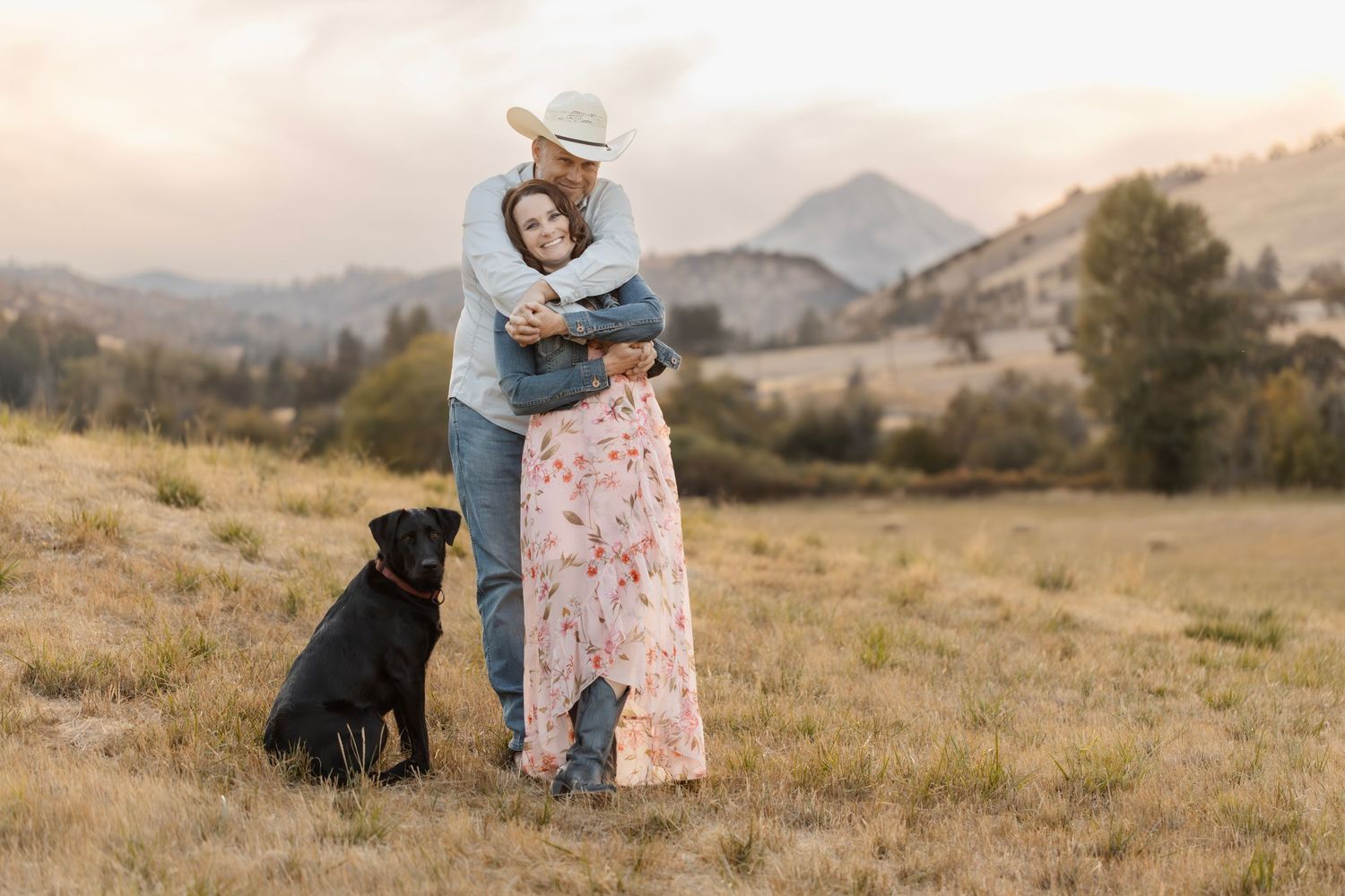 A couple and their black dog share a tender moment in a sunlit meadow with mountains in the background.