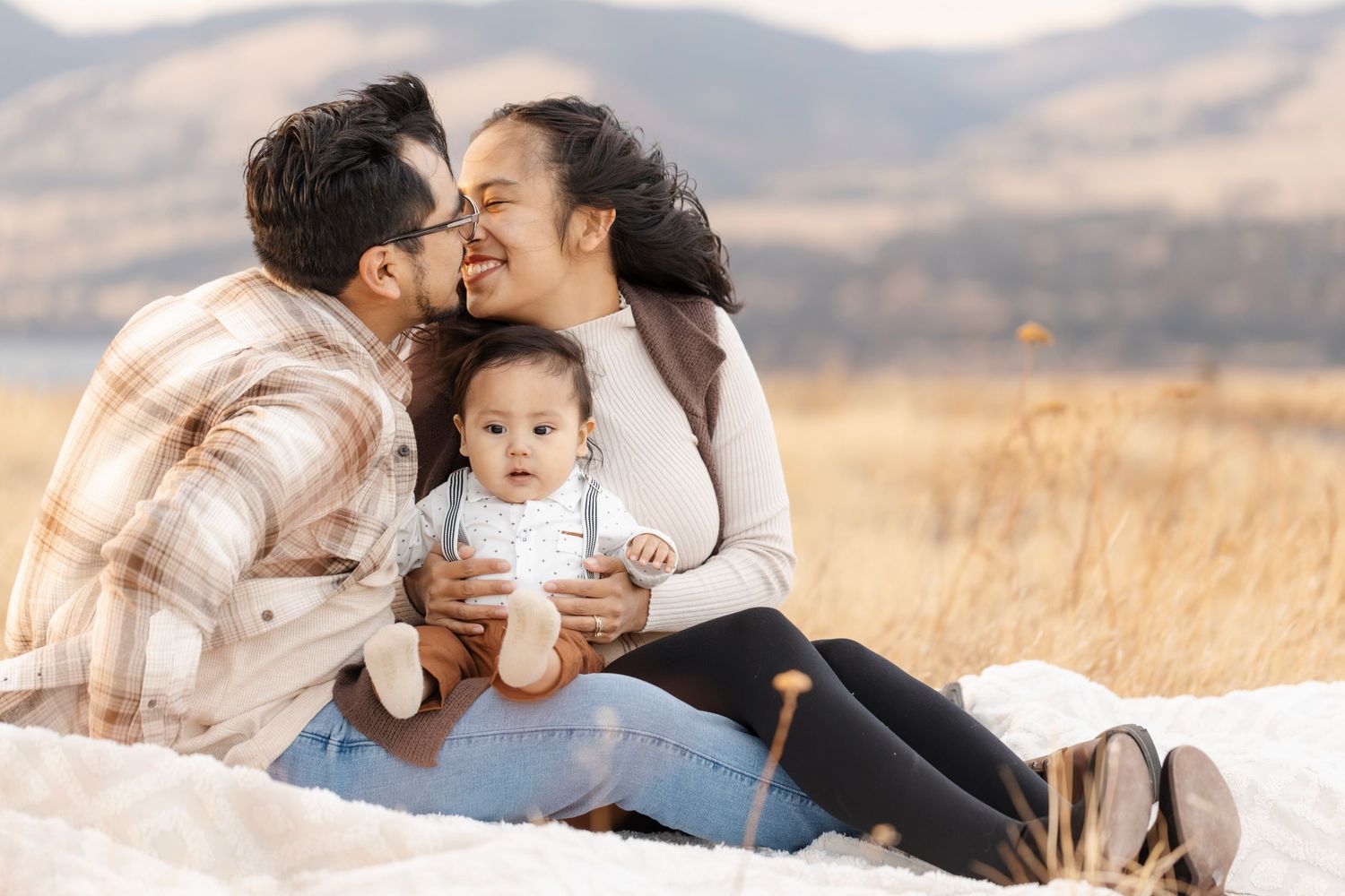 Intimate family moment shared on blanket in golden field with mountains behind.