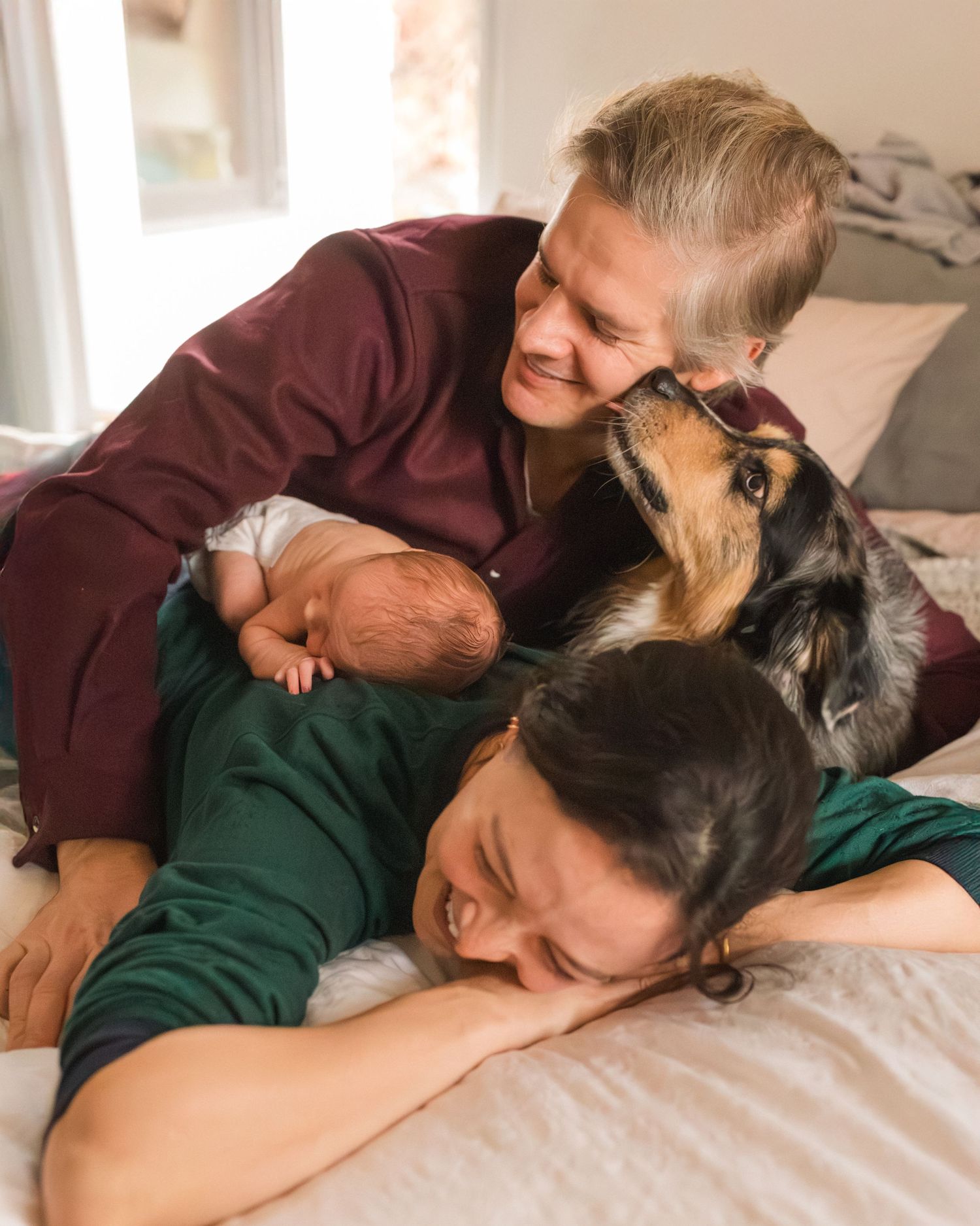 Two parents lying on a bed with their newborn baby while a friendly dog leans in to sniff.