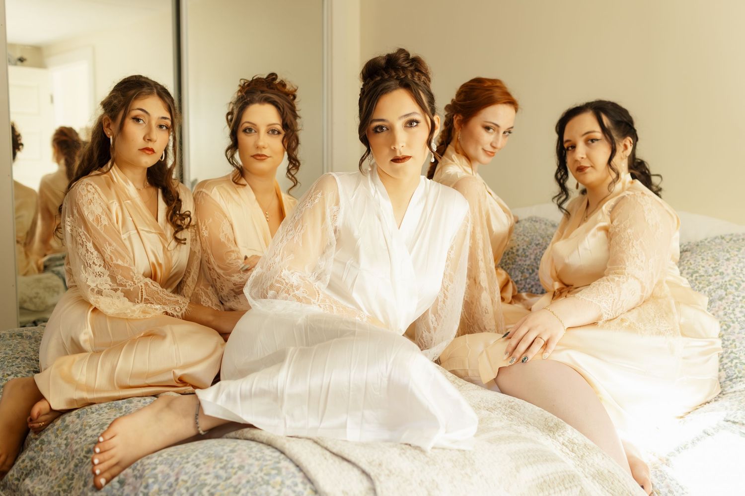 Five women in matching blush-colored robes lounging together on a bed during wedding preparations.