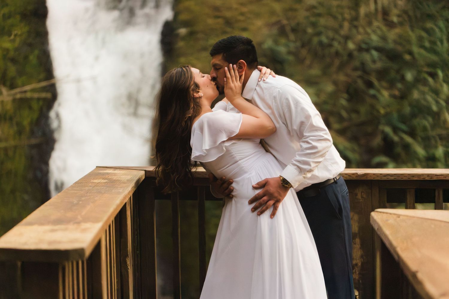 Couple sharing a romantic kiss on a wooden bridge in front of a cascading waterfall, bride in white dress.