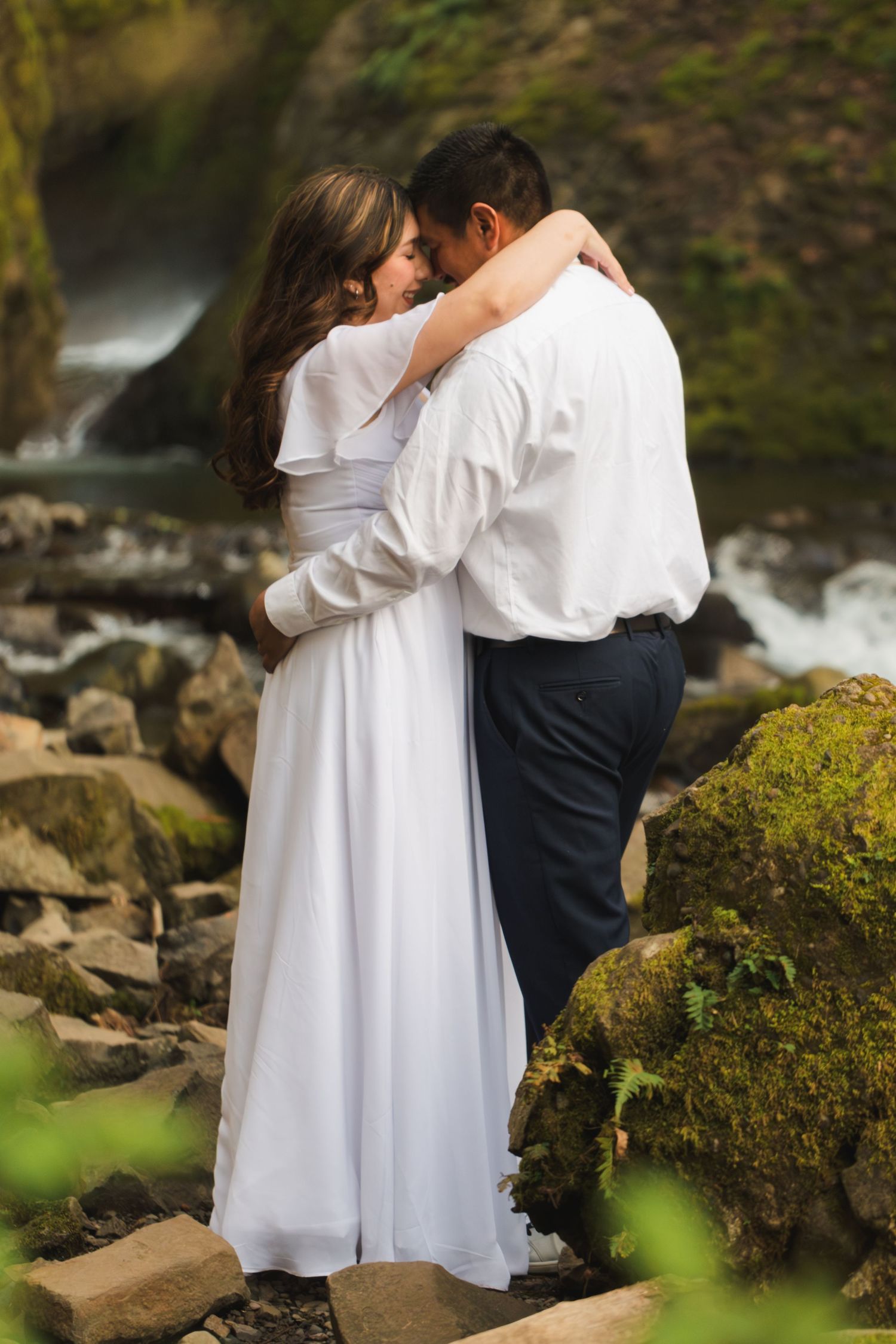 Couple embracing in white wedding attire on mossy rocks near a waterfall in a lush green forest.