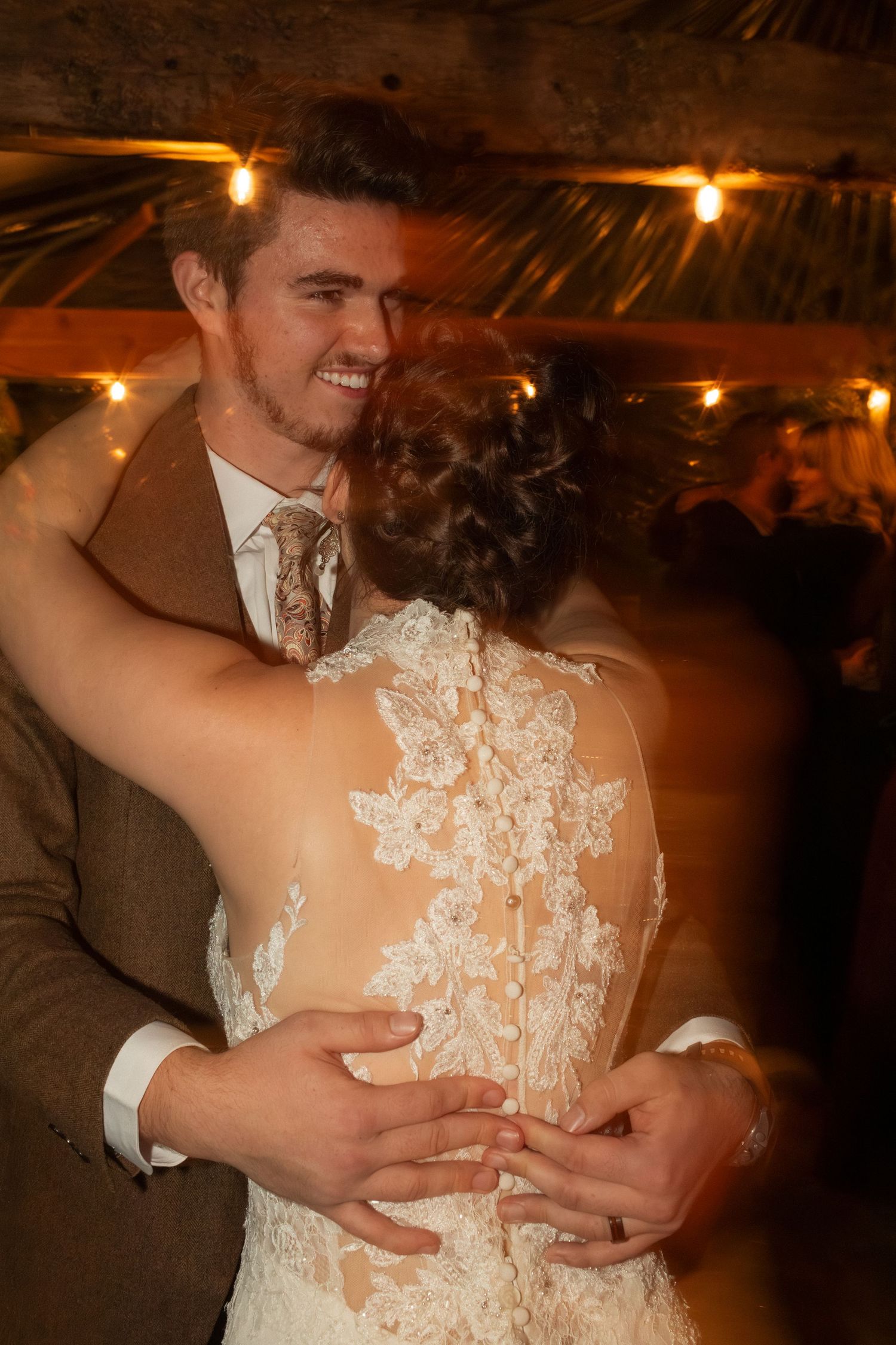 Couple embraces on dance floor under warm lighting showing intricate lace back wedding dress detail.