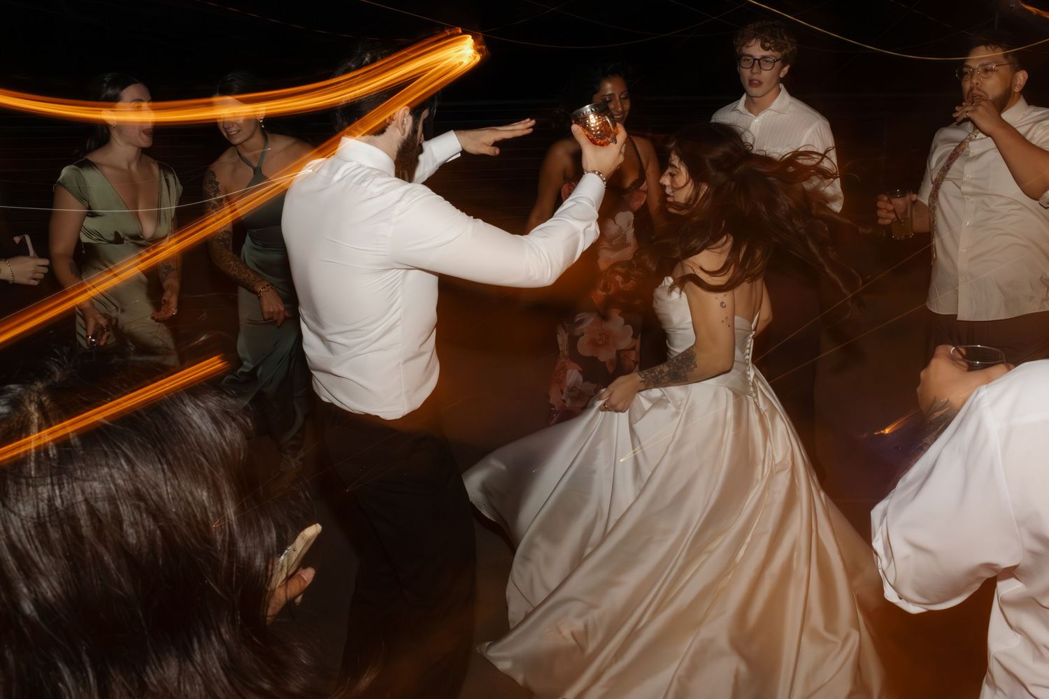 Wedding reception guests dance and celebrate on a dance floor with glowing orange lights overhead.