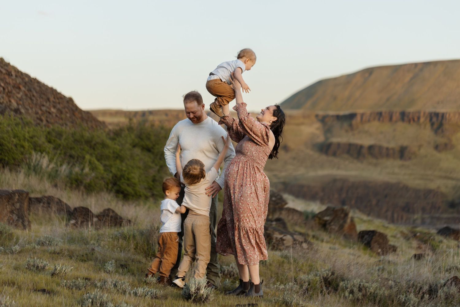 Family of five posing outdoors in a scenic landscape, mother lifting baby overhead while father and children stand nearby.