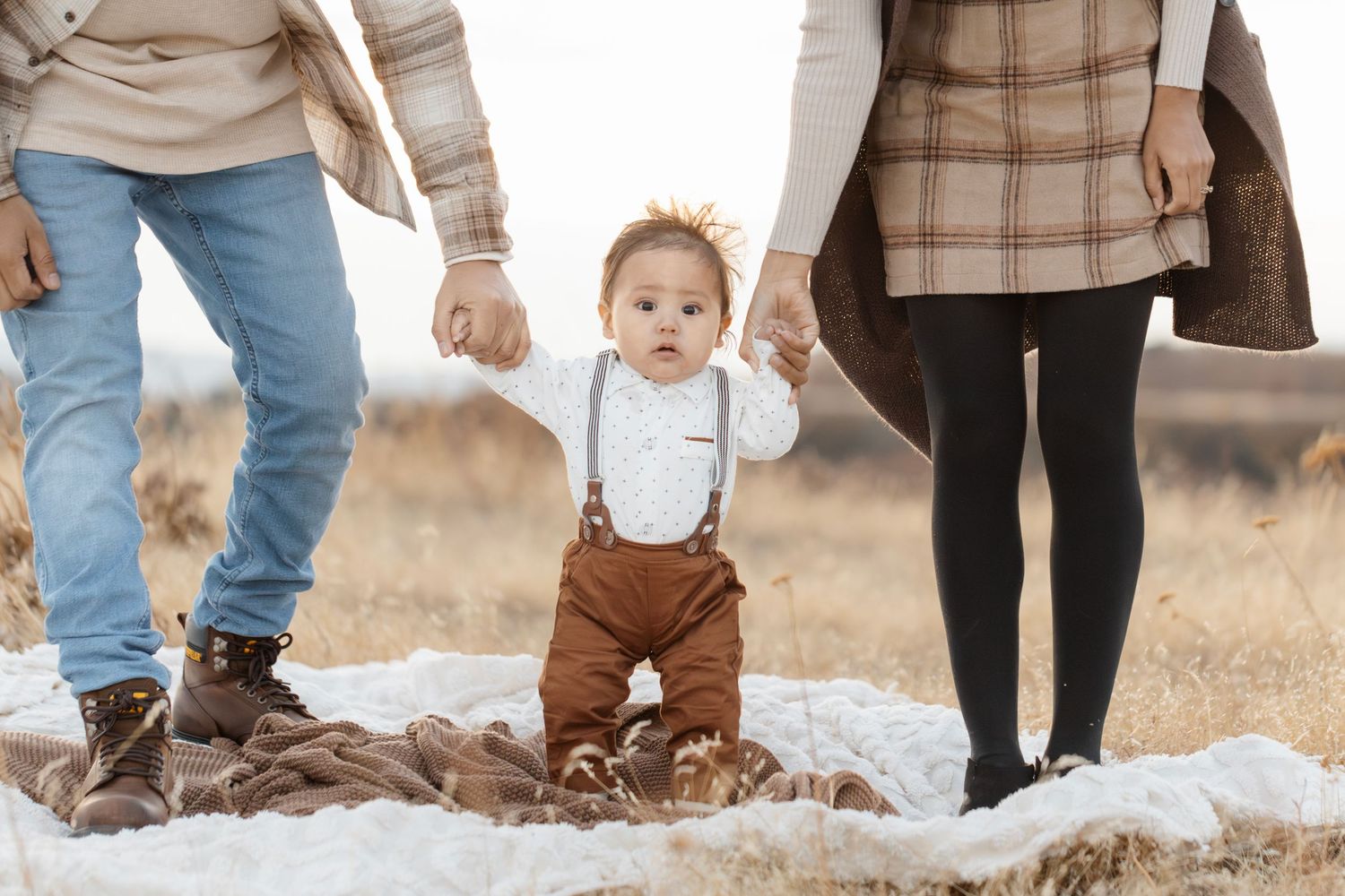 Small child in brown overalls takes first steps between parents on snowy ground.