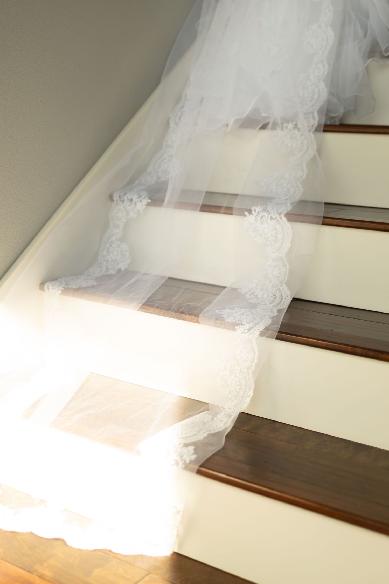 Sunlight streams through a window onto white lace fabric draped over wooden stairs creating ethereal shadows and patterns.