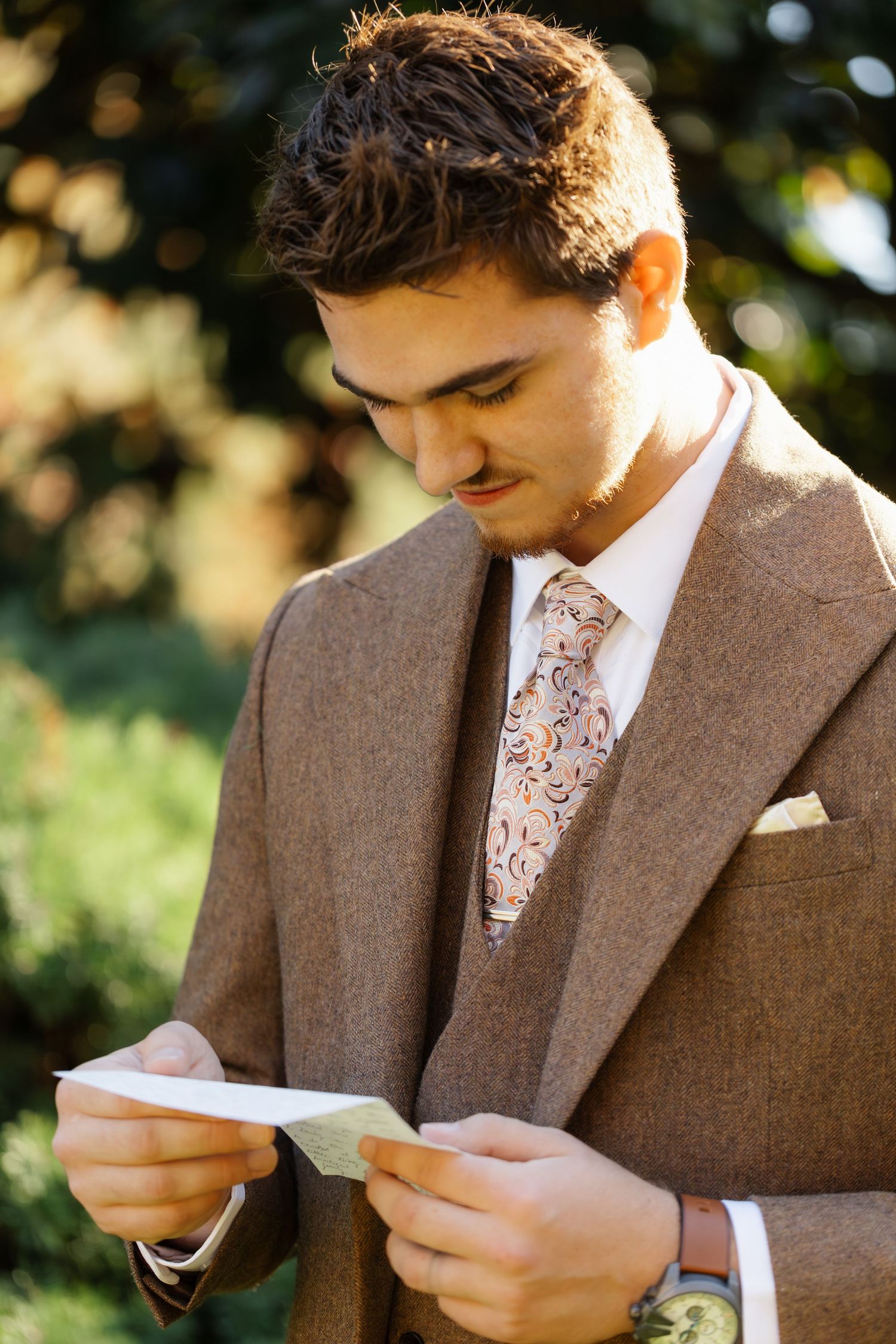 Groom in brown tweed suit reads an emotional letter outdoors.