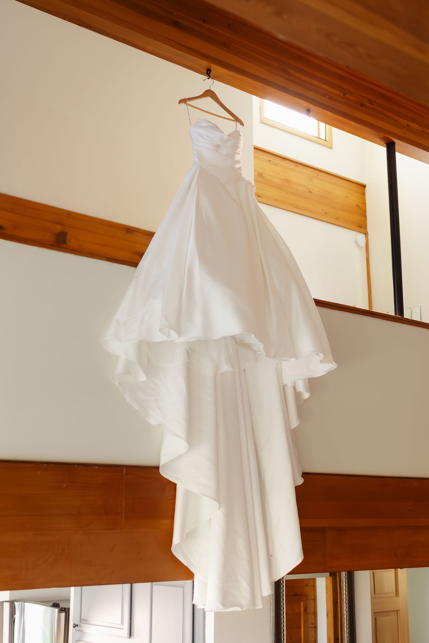 White wedding dress hangs elegantly from wooden beams in natural lighting creating an ethereal display.