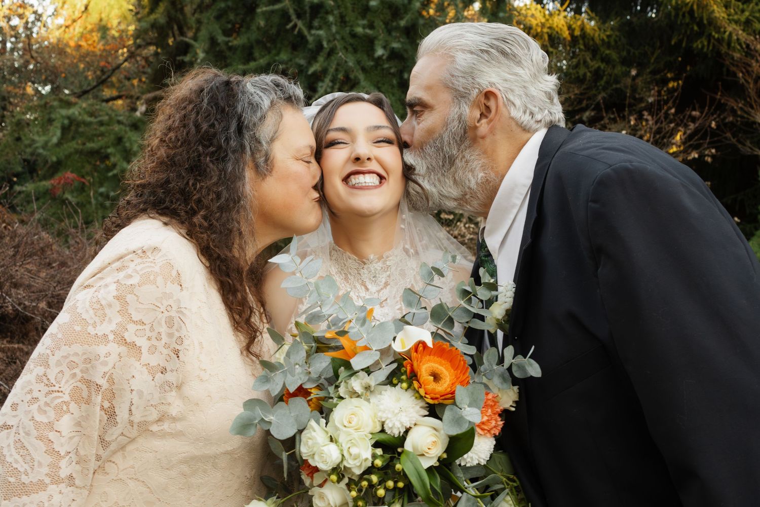 Bride smiling joyfully as parents kiss her cheeks, holding orange and white floral bouquet outdoors.