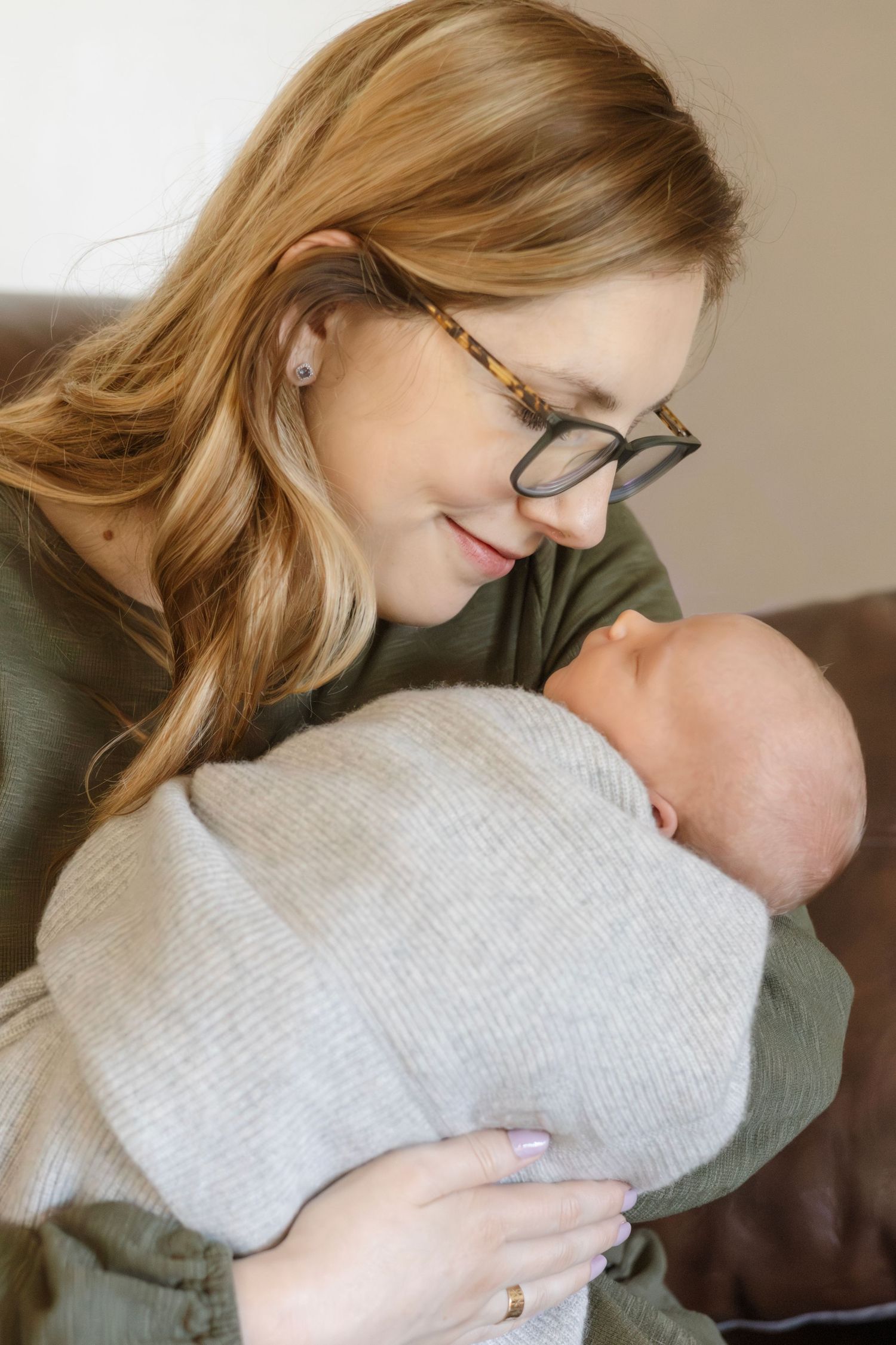 Mother in olive sweater tenderly holds sleeping newborn against her chest.