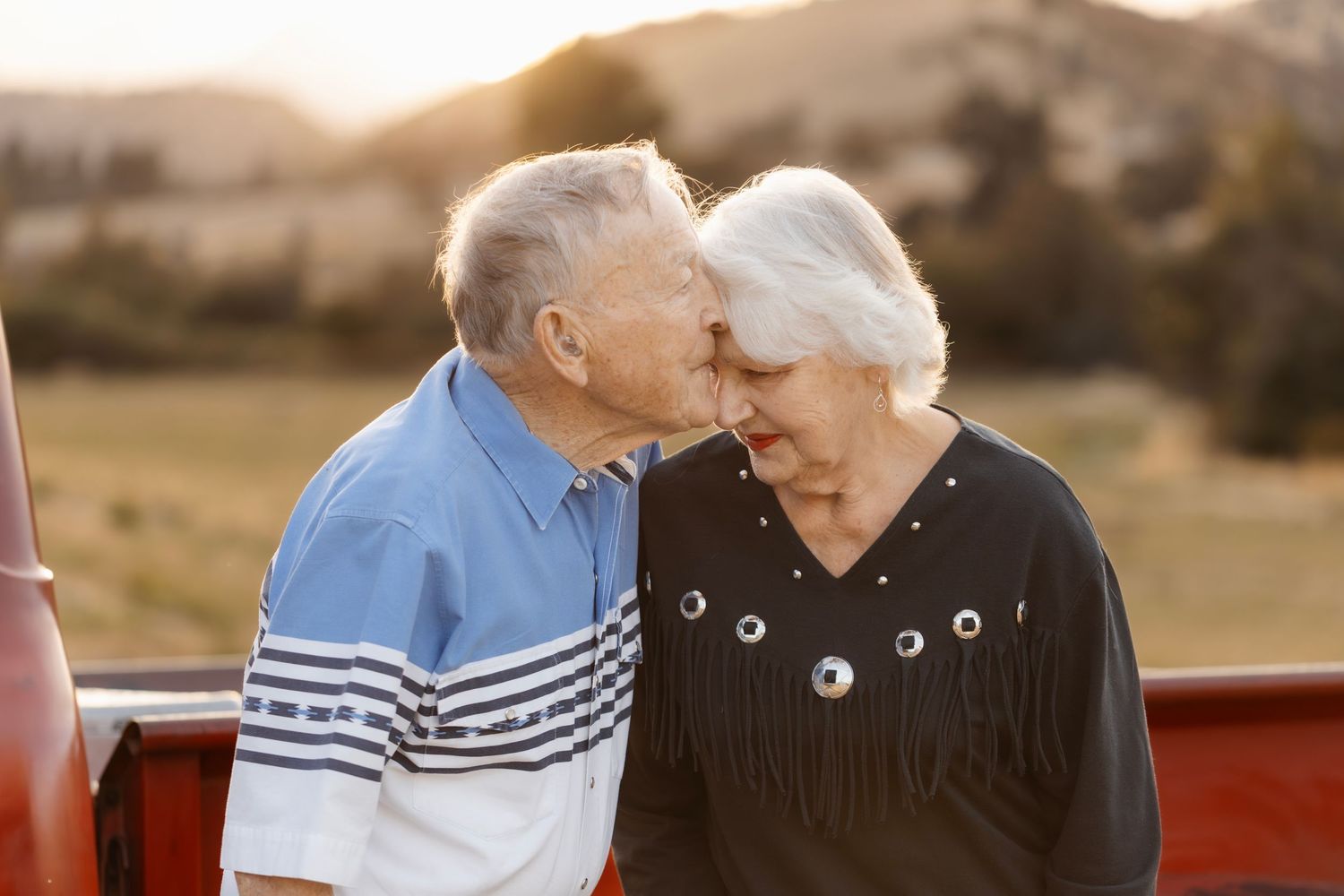 An elderly couple shares a tender moment beside a vintage red truck at sunset.