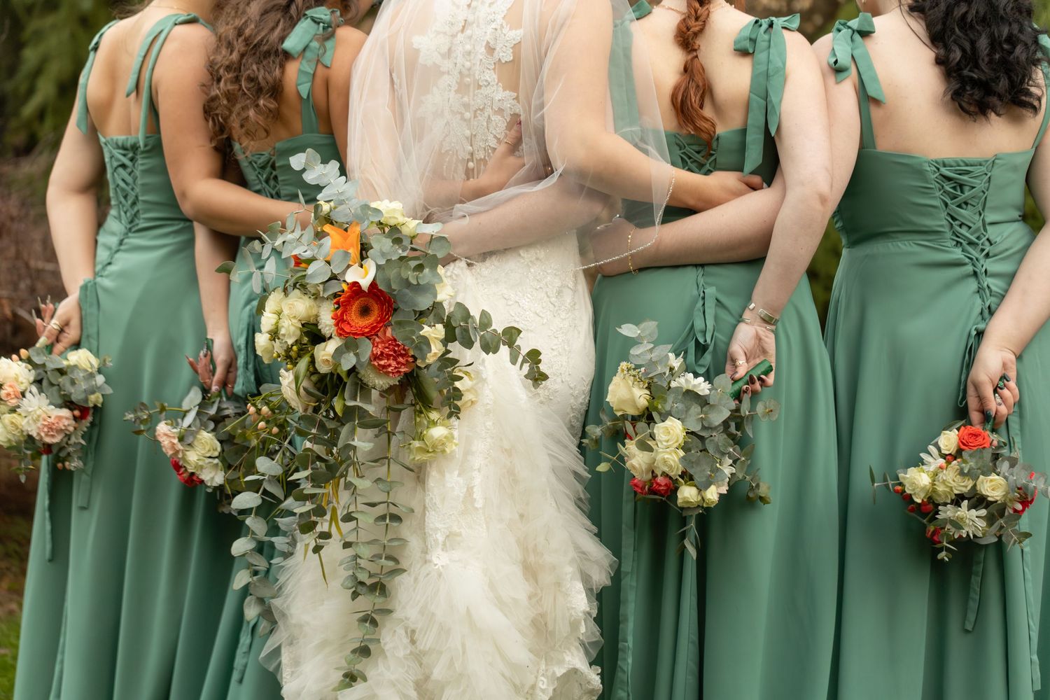 Bridesmaids in sage green dresses hold colorful autumn bouquets at an outdoor wedding ceremony.