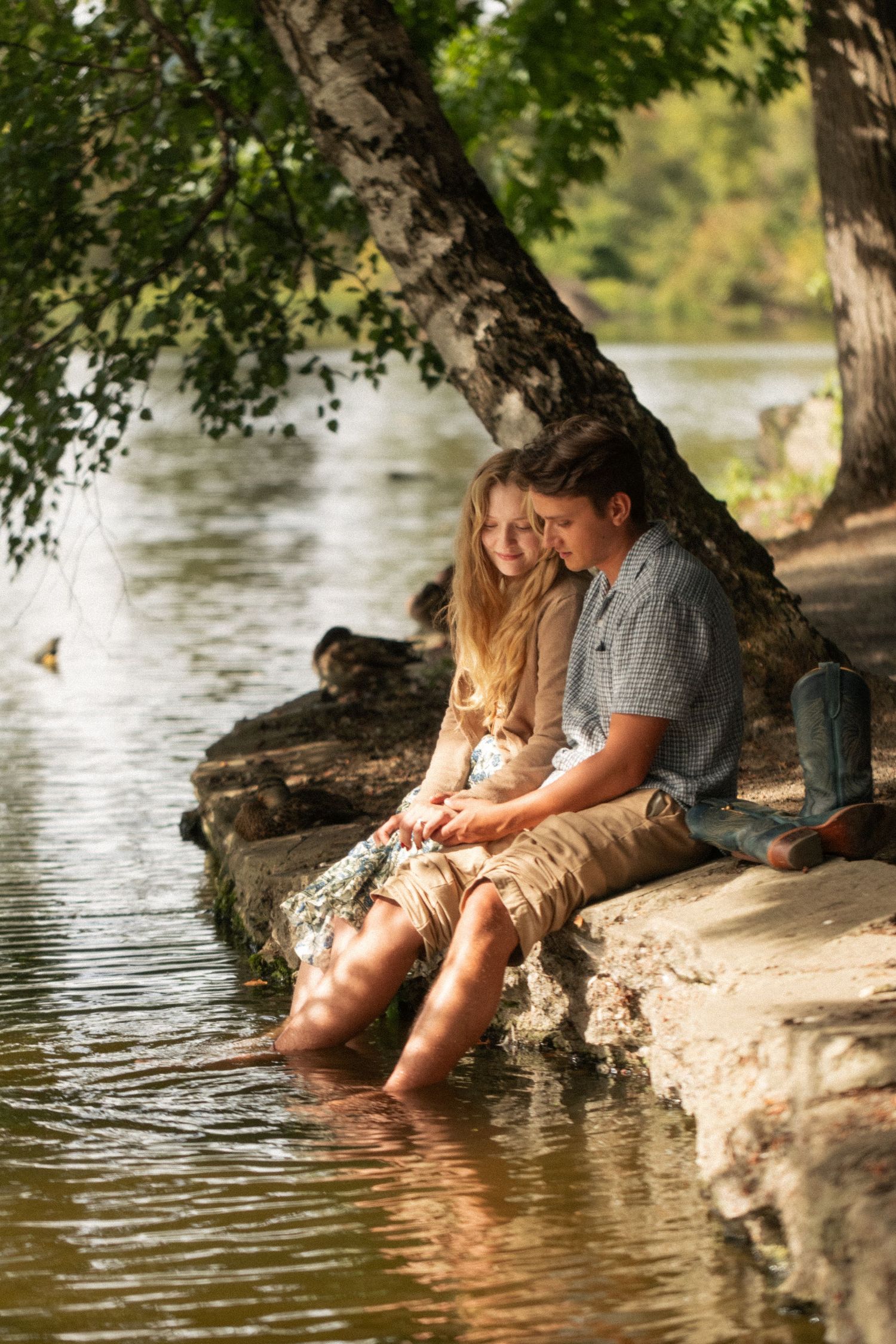 Young couple sits close together on a rocky shore beneath a leaning tree, dipping their bare feet in calm lake water.