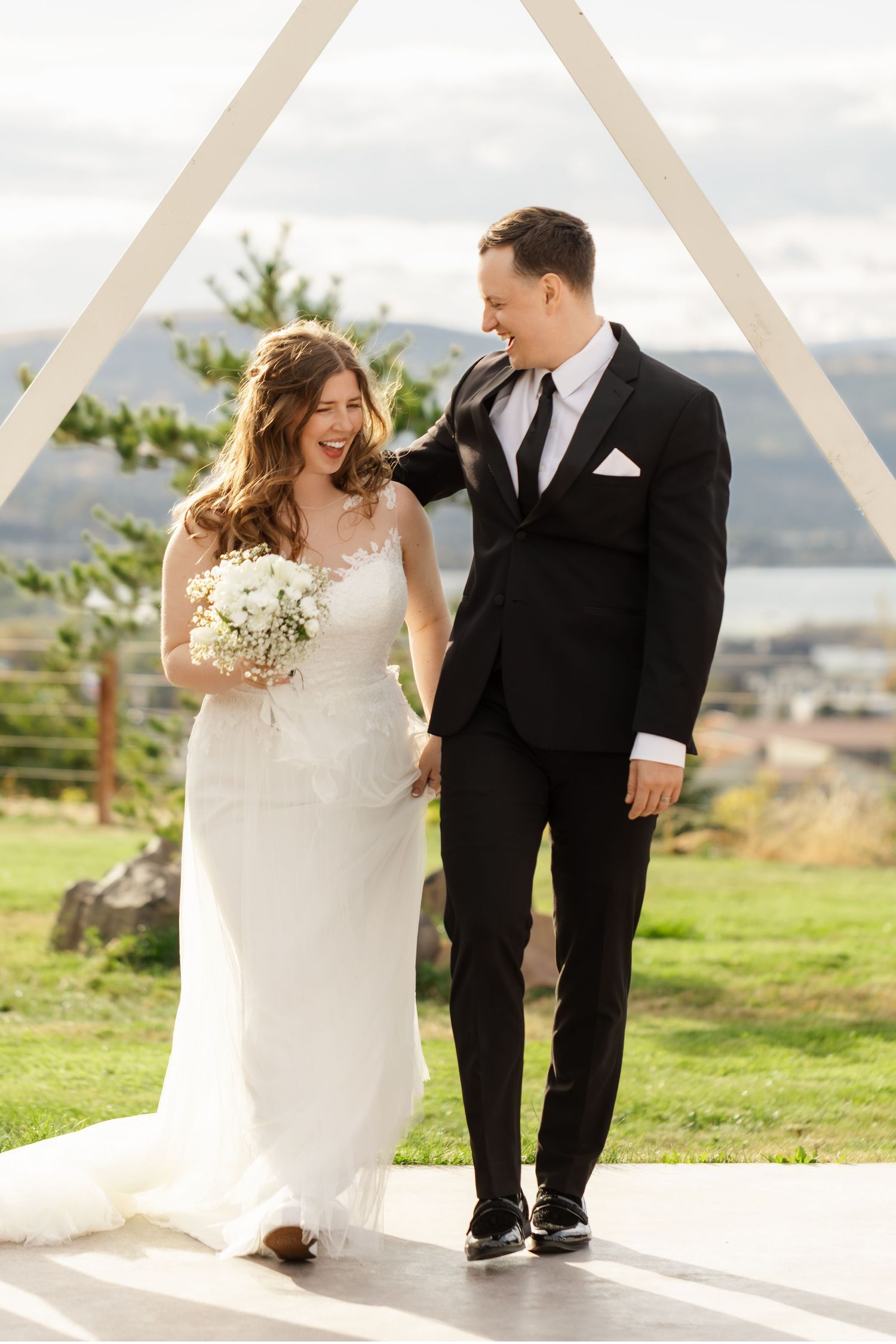 Couple in wedding attire walks together and laughs on a scenic hilltop path during their special day.