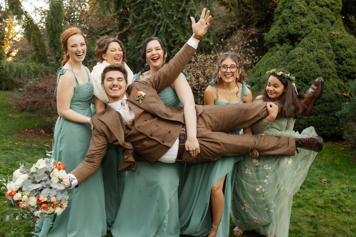 Groom carried by laughing bridesmaids in sage green dresses at outdoor wedding celebration.