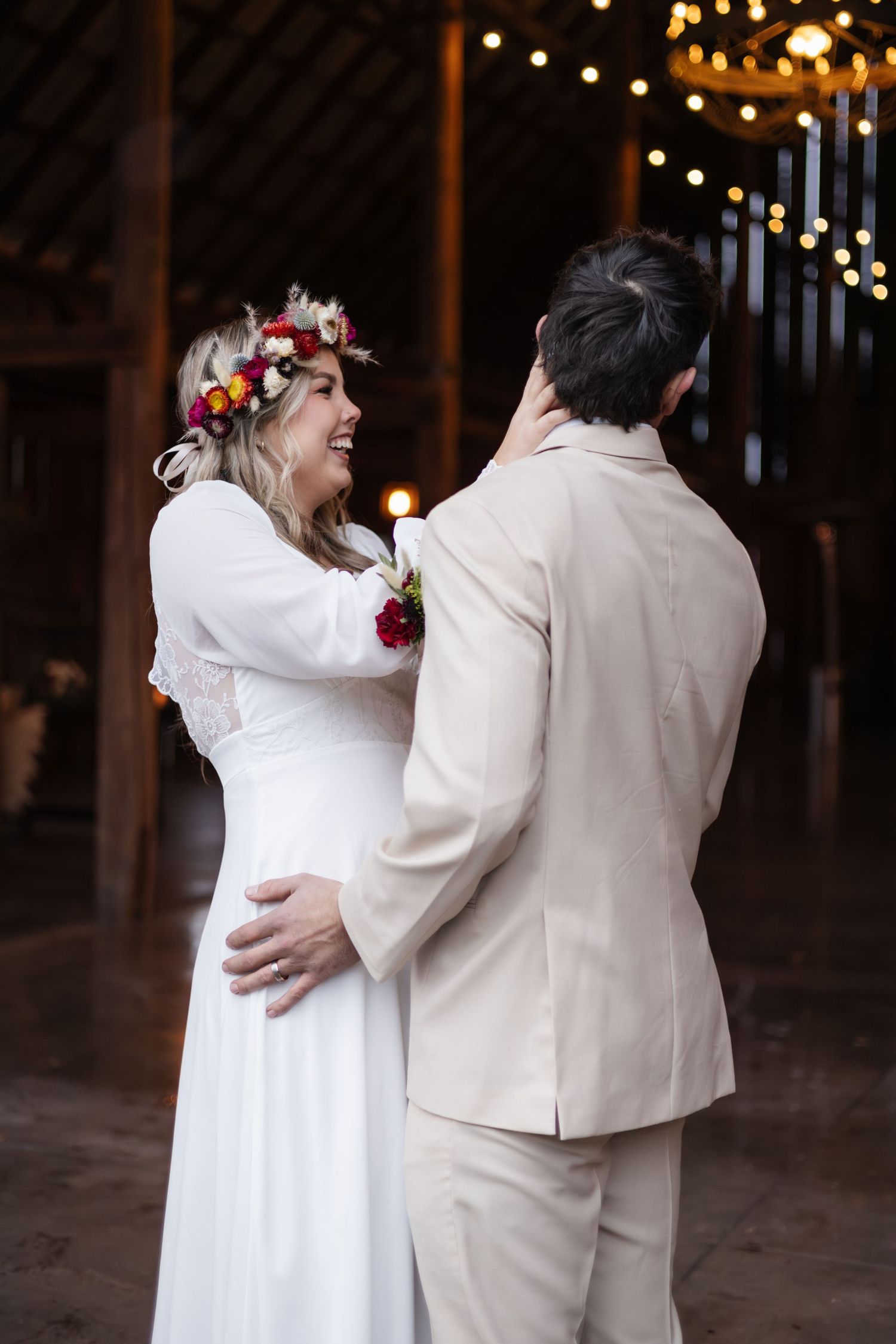 Newlyweds share intimate moment, bride in white dress and floral crown, groom in light suit under string lights.