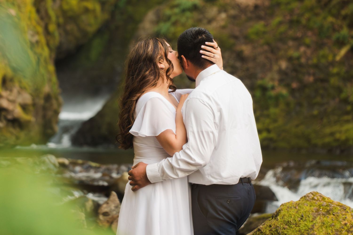 Couple sharing a romantic kiss near a waterfall, woman in white dress embracing man in white shirt outdoors.