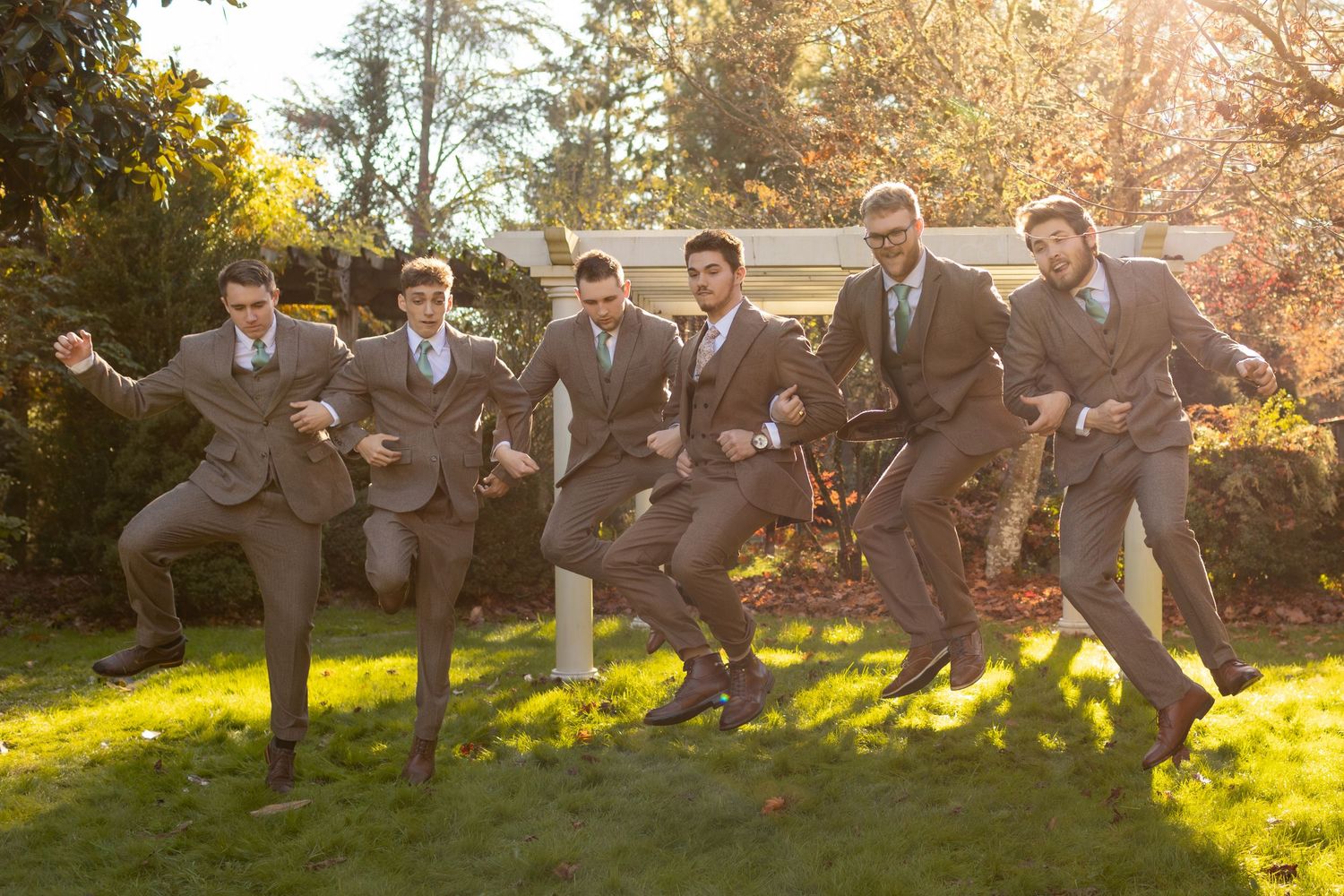 Six groomsmen in gray suits striking fun dynamic poses on green grass with sunlit trees in the background.