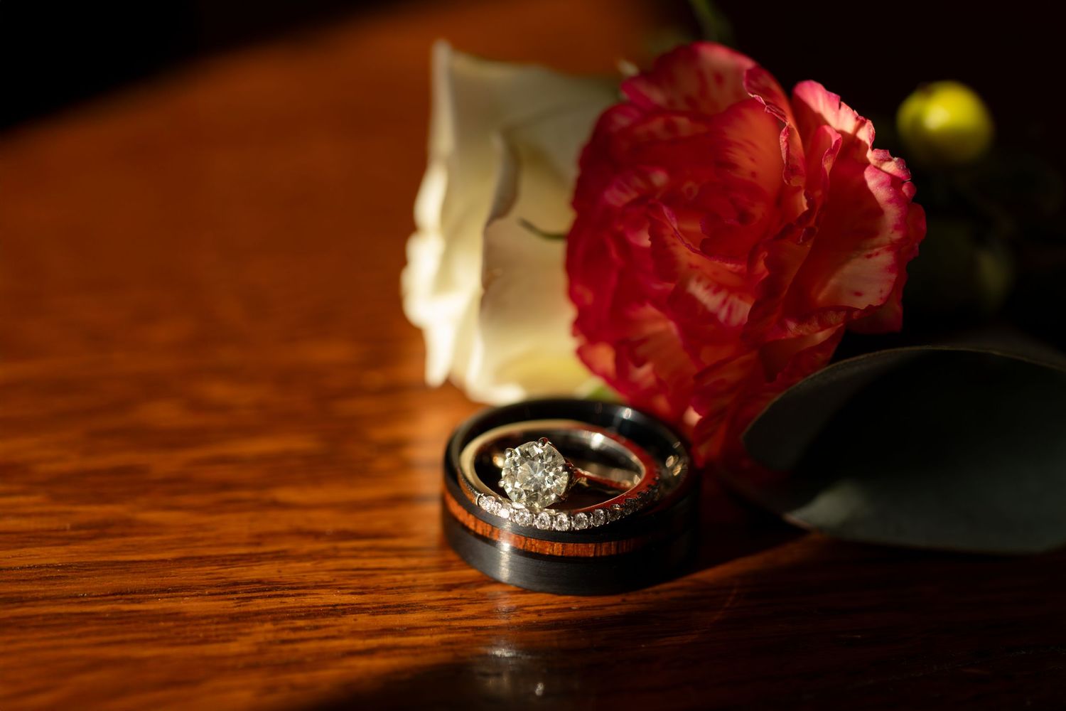 Red carnation and wedding ring detail shot on dark wooden surface with dramatic lighting.