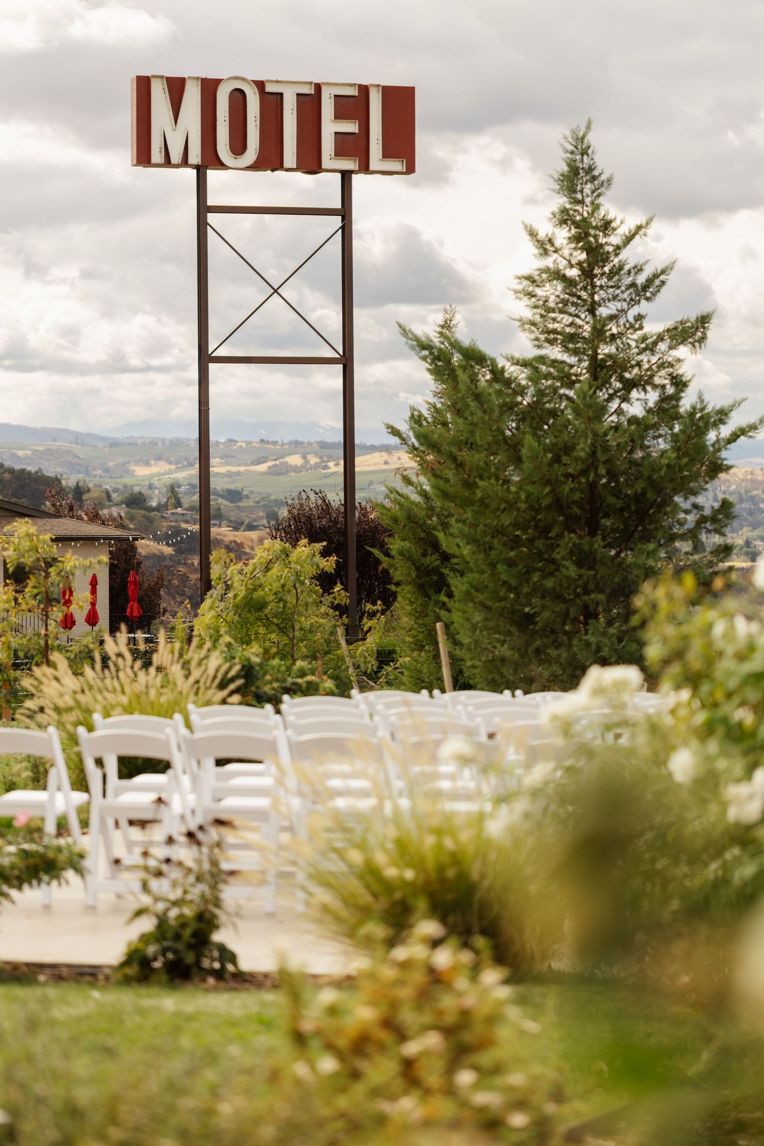 White chairs arranged outdoors for a wedding ceremony with a motel sign and mountains in the background.