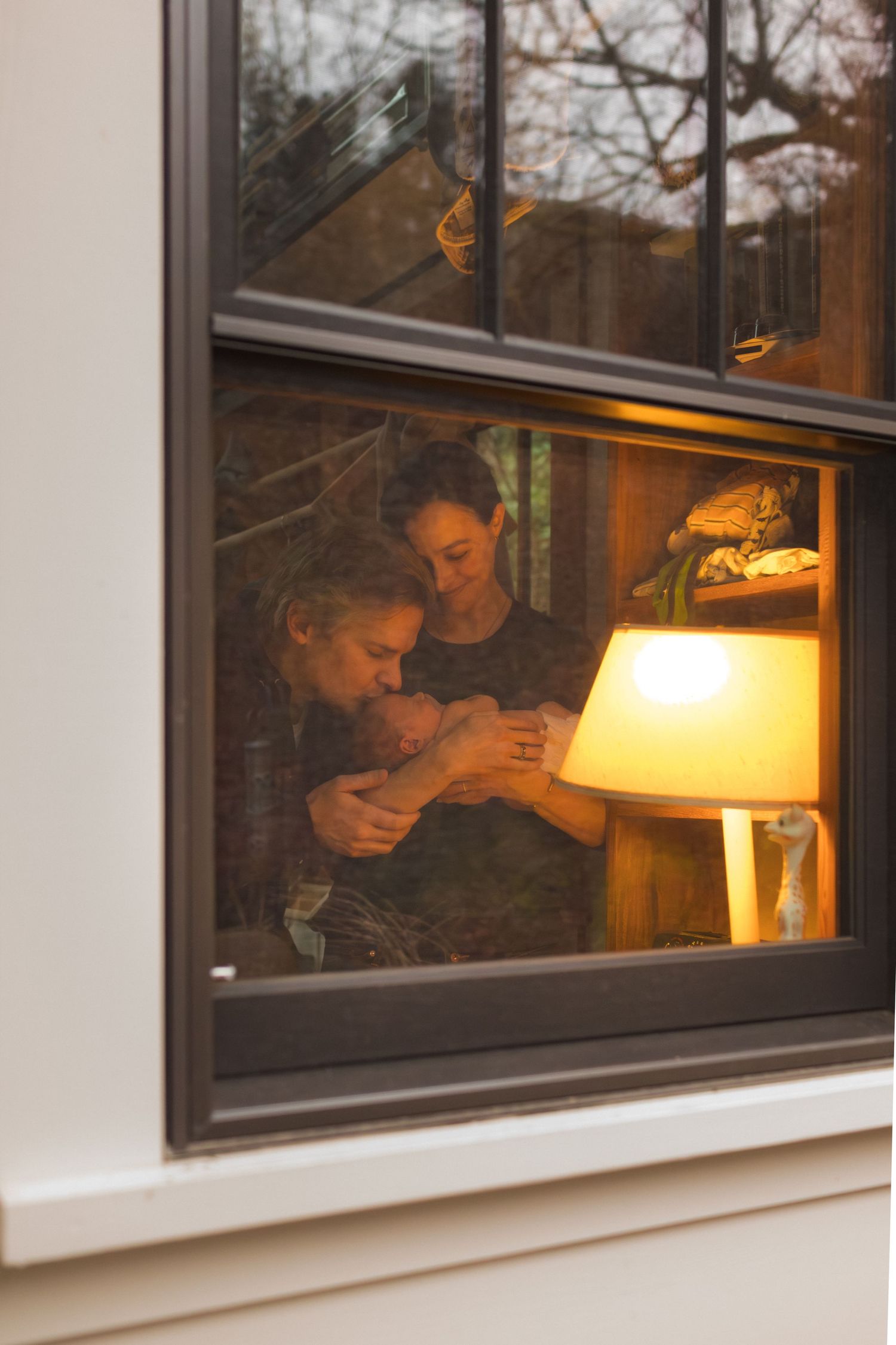 Two people sharing a tender moment with a newborn baby by a warm lamp, viewed through a window at dusk.