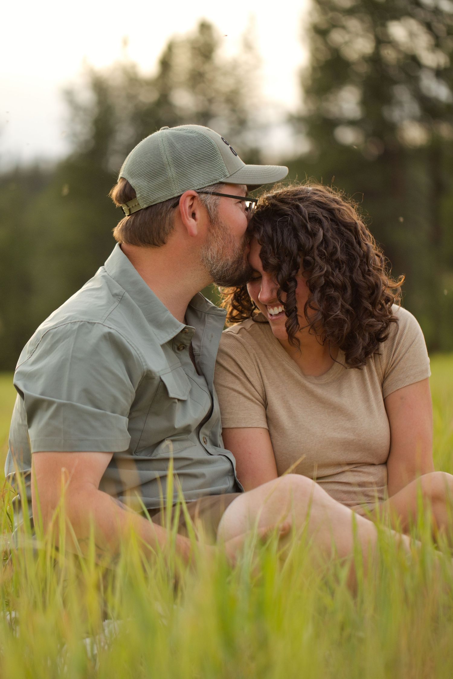 A couple sits in tall grass, sharing a tender moment as the man kisses the smiling woman on the forehead outdoors.