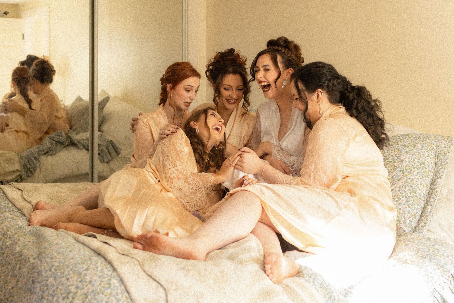 Group of friends in soft peach dresses lounging together on white bedding during a relaxed photoshoot.