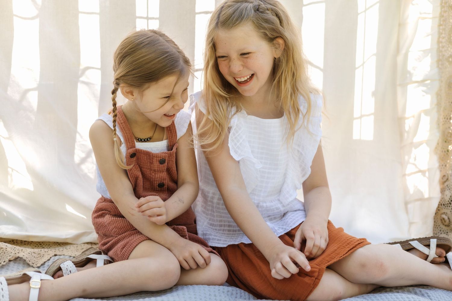 Two children sitting together on a blanket share a joyful laugh in soft natural lighting.