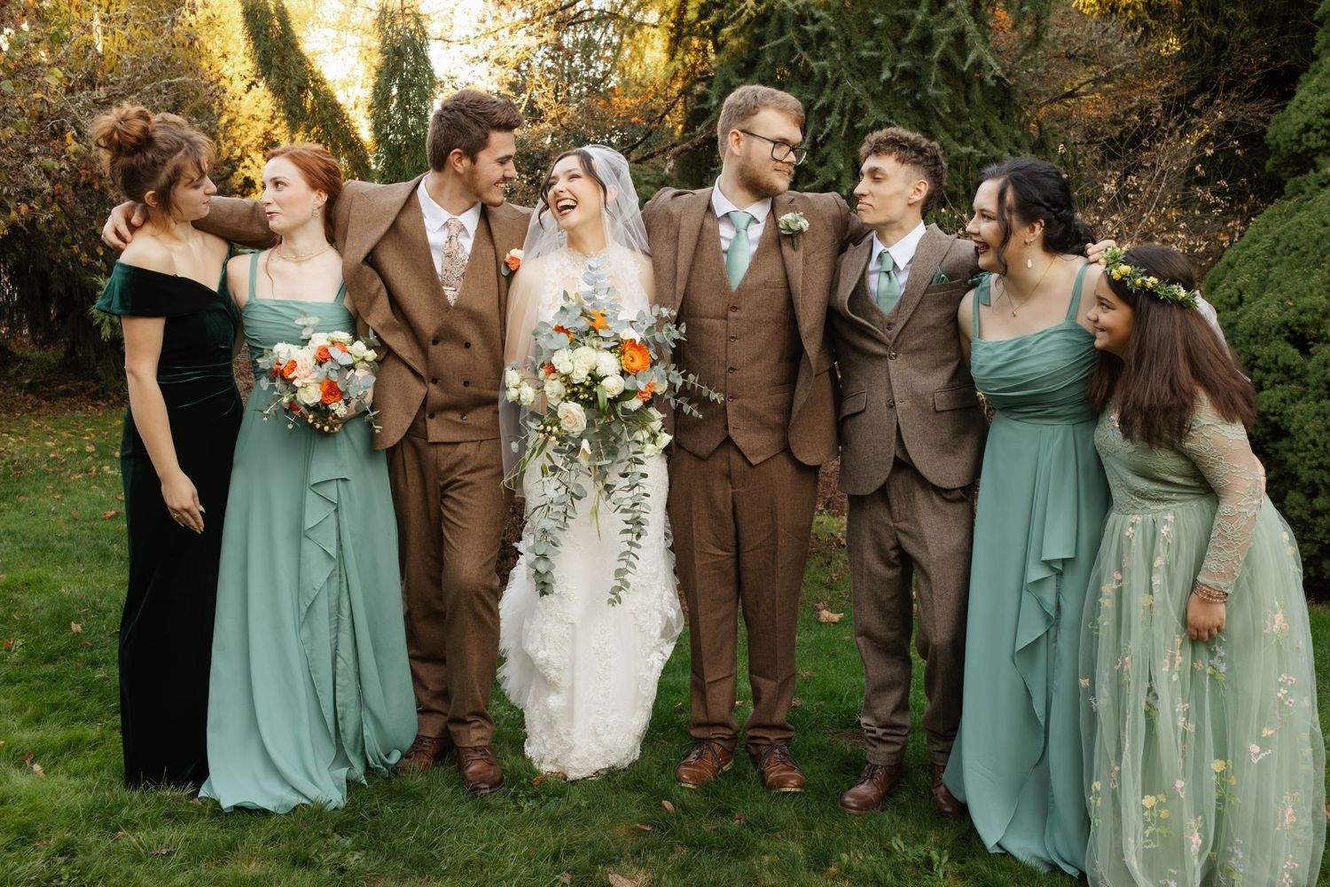 Wedding party in brown suits and sage green dresses posing outdoors, bride holding lush floral bouquet.