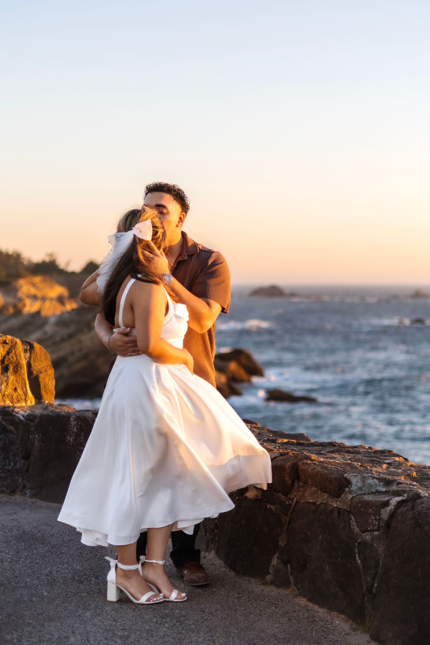 A romantic sunset photoshoot on rocky coastal cliffs with a white dress against an orange sky.
