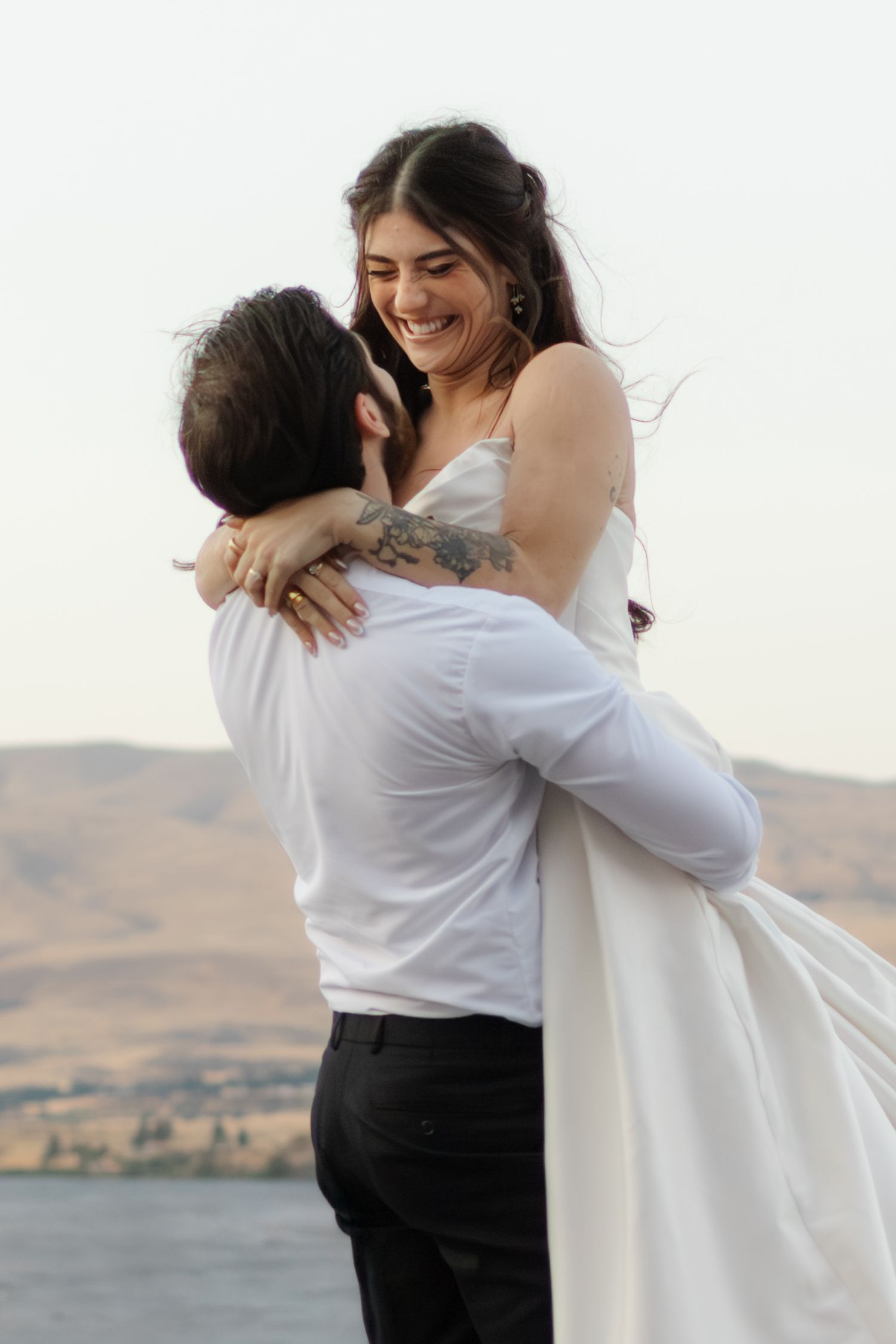 A couple embraces in white attire against a scenic desert backdrop during their wedding celebration.
