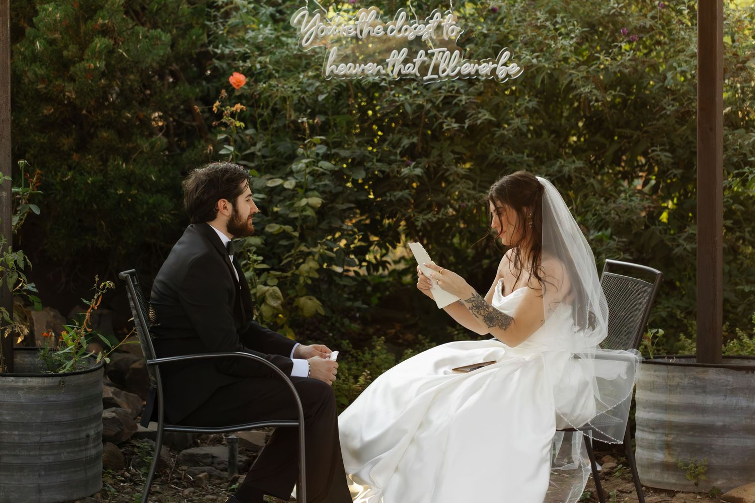 Bride and groom in wedding attire sit outdoors reading vows, surrounded by lush greenery.