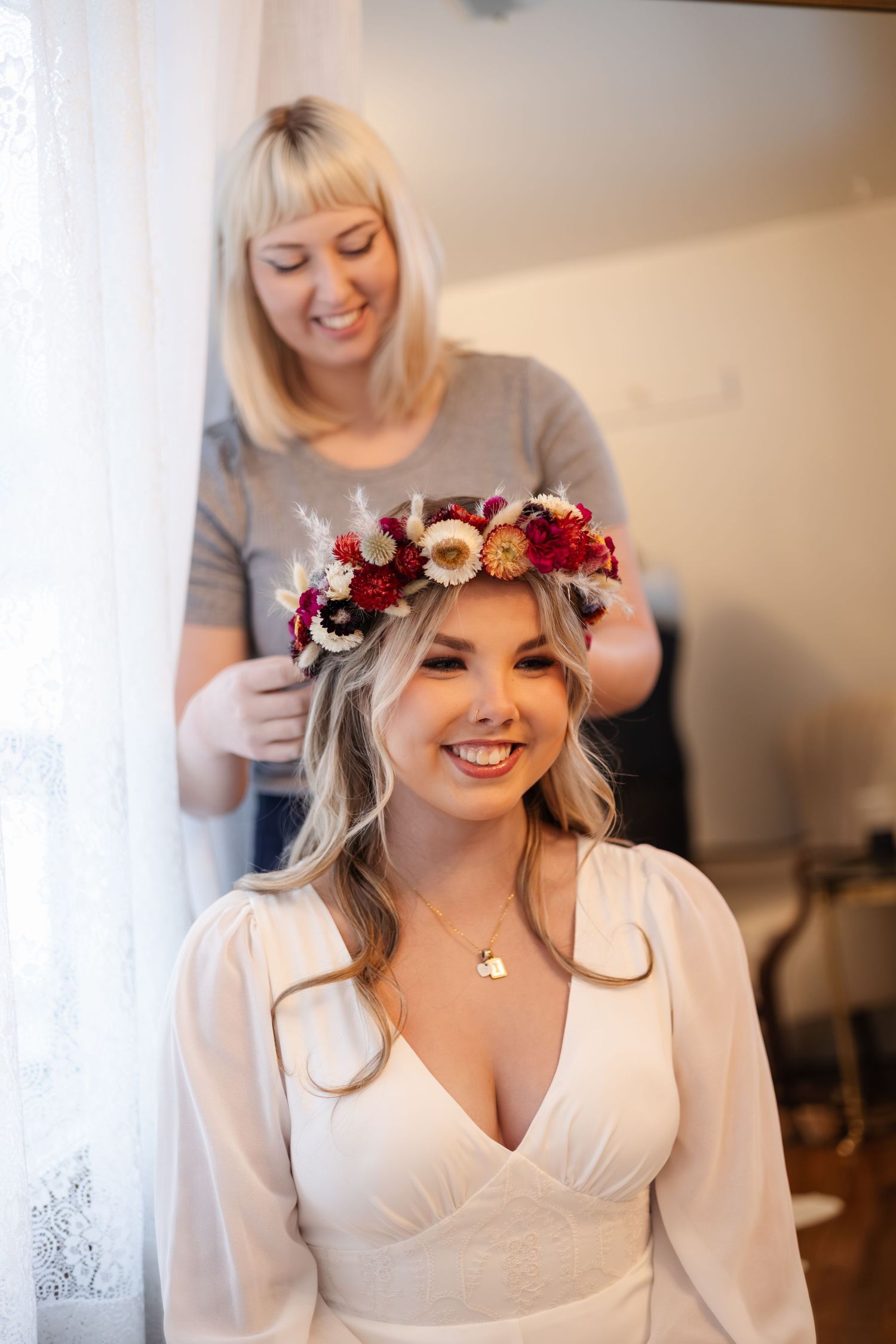 A floral crown being placed on a woman wearing a white dress during wedding preparations.