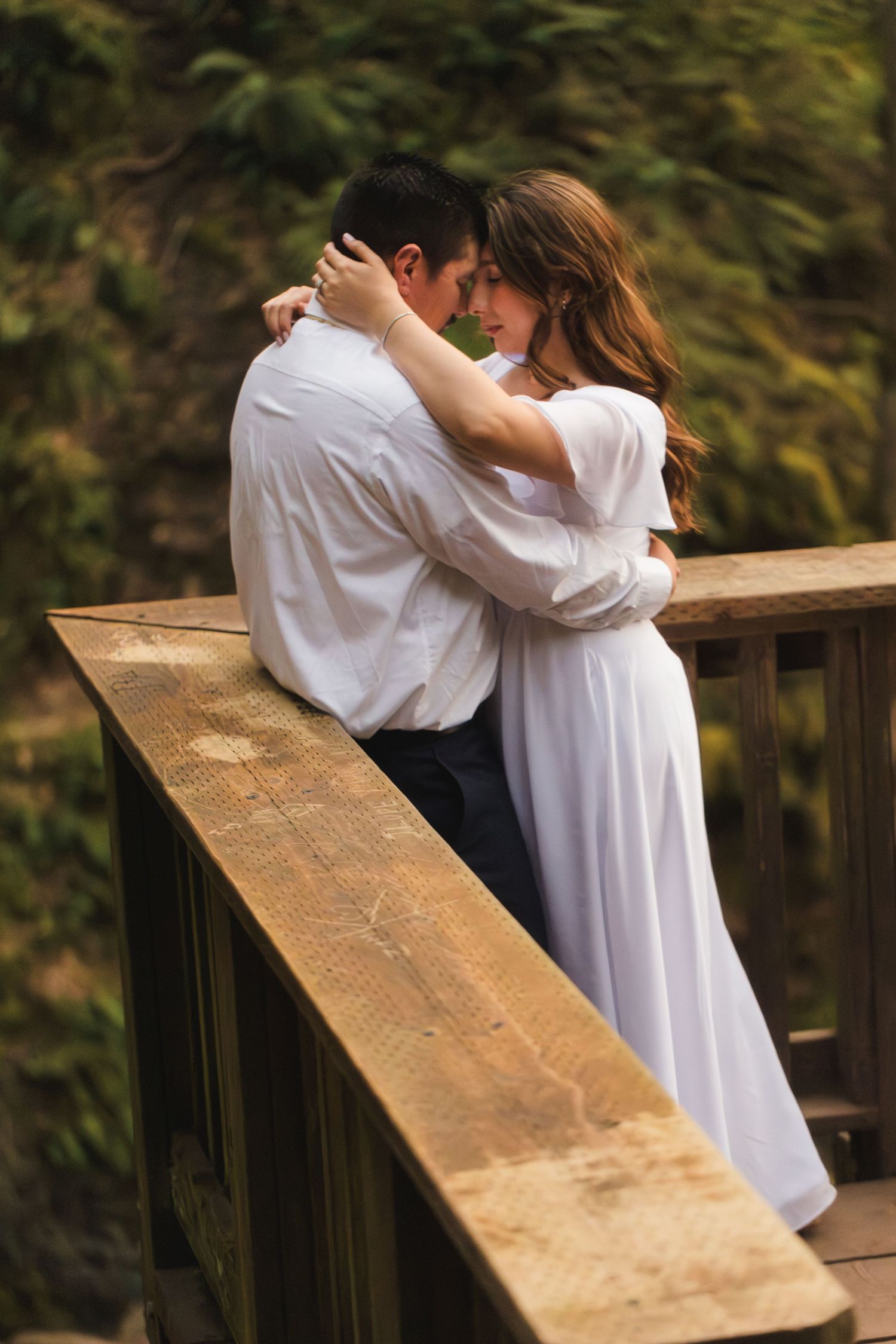 Couple embracing romantically on a wooden bridge, woman in white dress with arms around mans neck, lush green background.