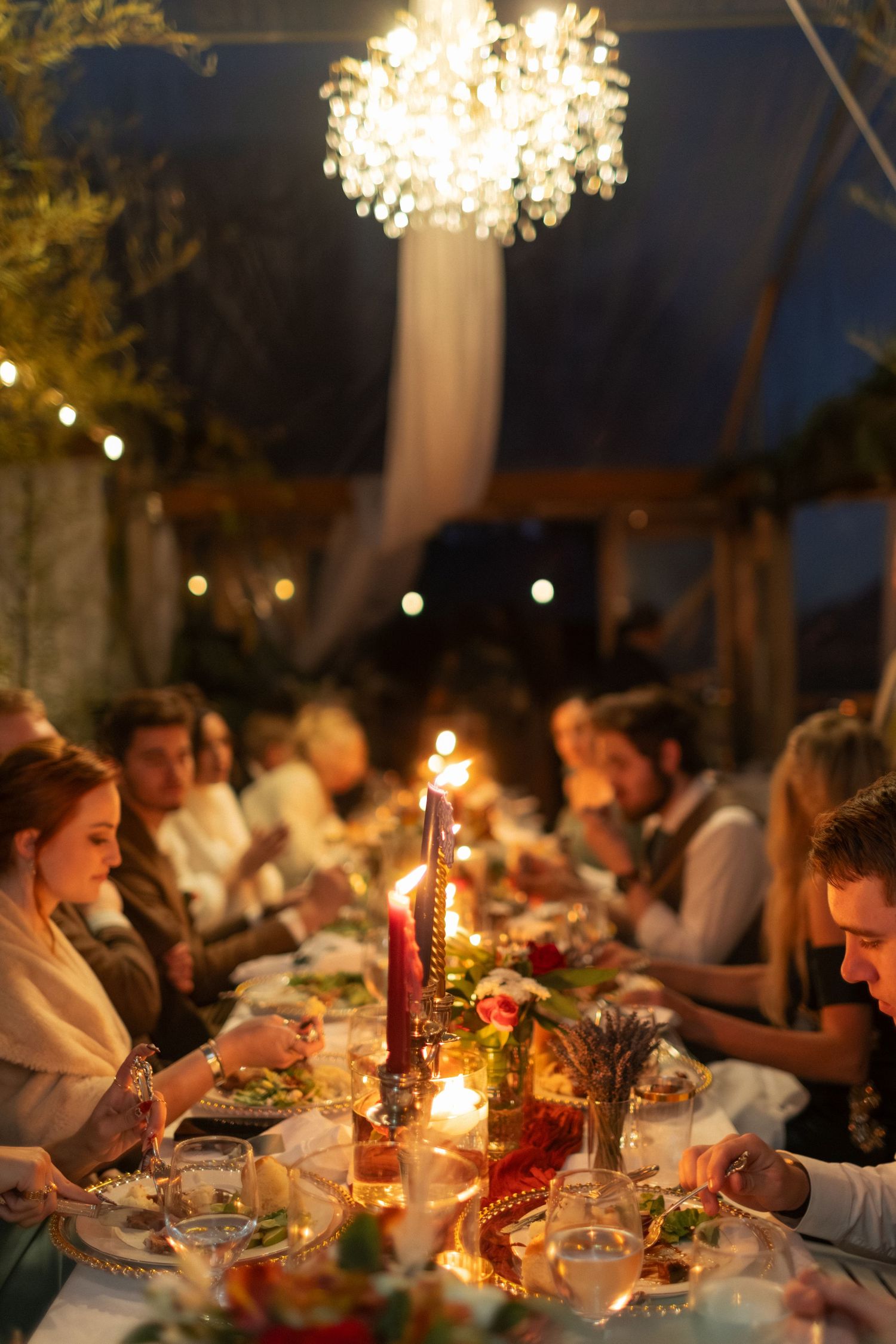 Candlelit wedding reception dinner under a crystal chandelier with guests dining at a long table in an outdoor setting.